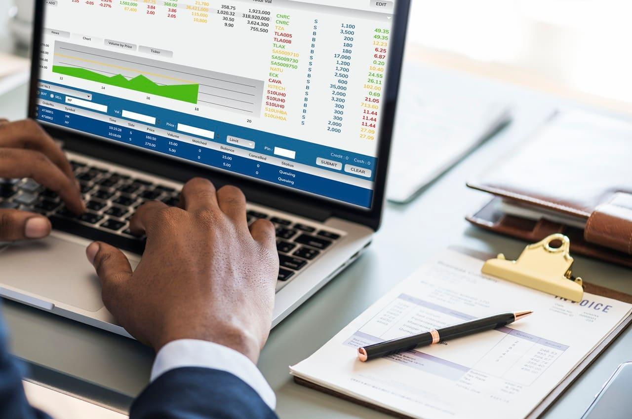 Pessoa analisando gráficos de ações de trade finance em um notebook. Há relatórios e documentos ao seu redor.