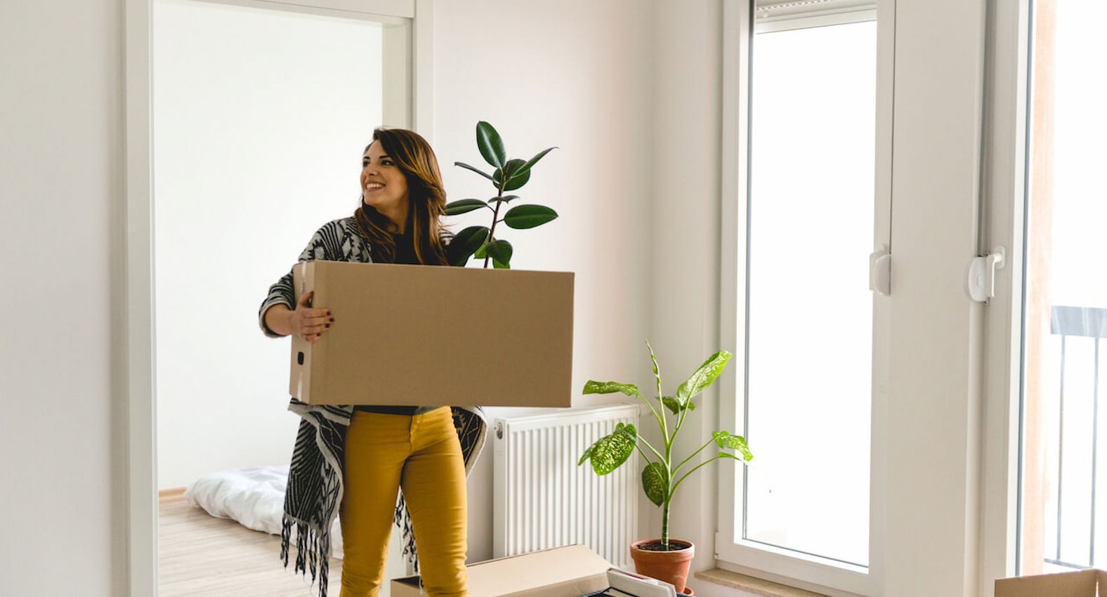 A imagem mostra uma mulher carregando uma caixa de papelão, se mudando para seu apartamento novo.