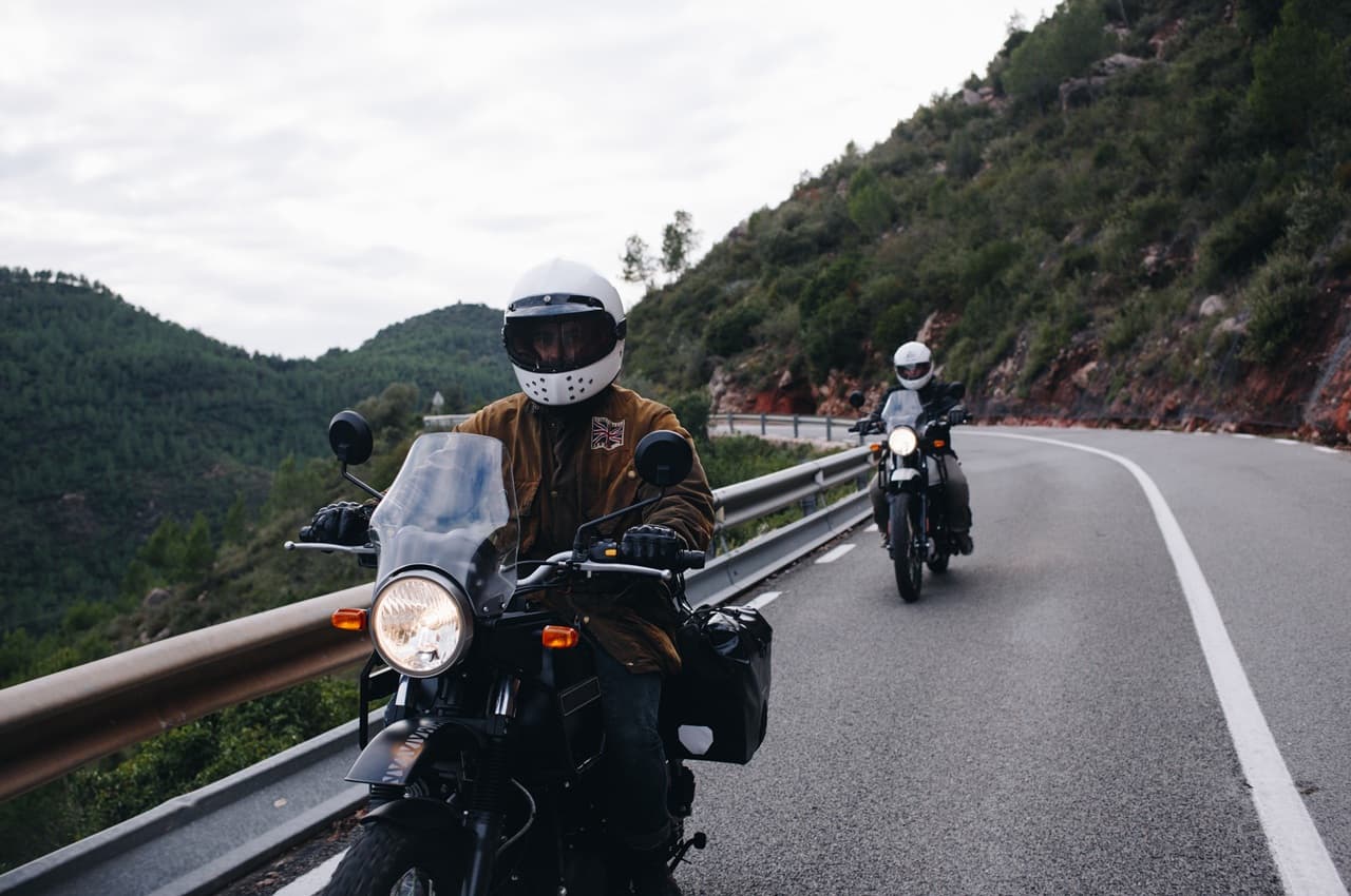 Duas pessoas pilotando motos de viagem em uma estrada sinuosa, com paisagem de montanhas e árvores ao fundo, sob céu parcialmente nublado.
