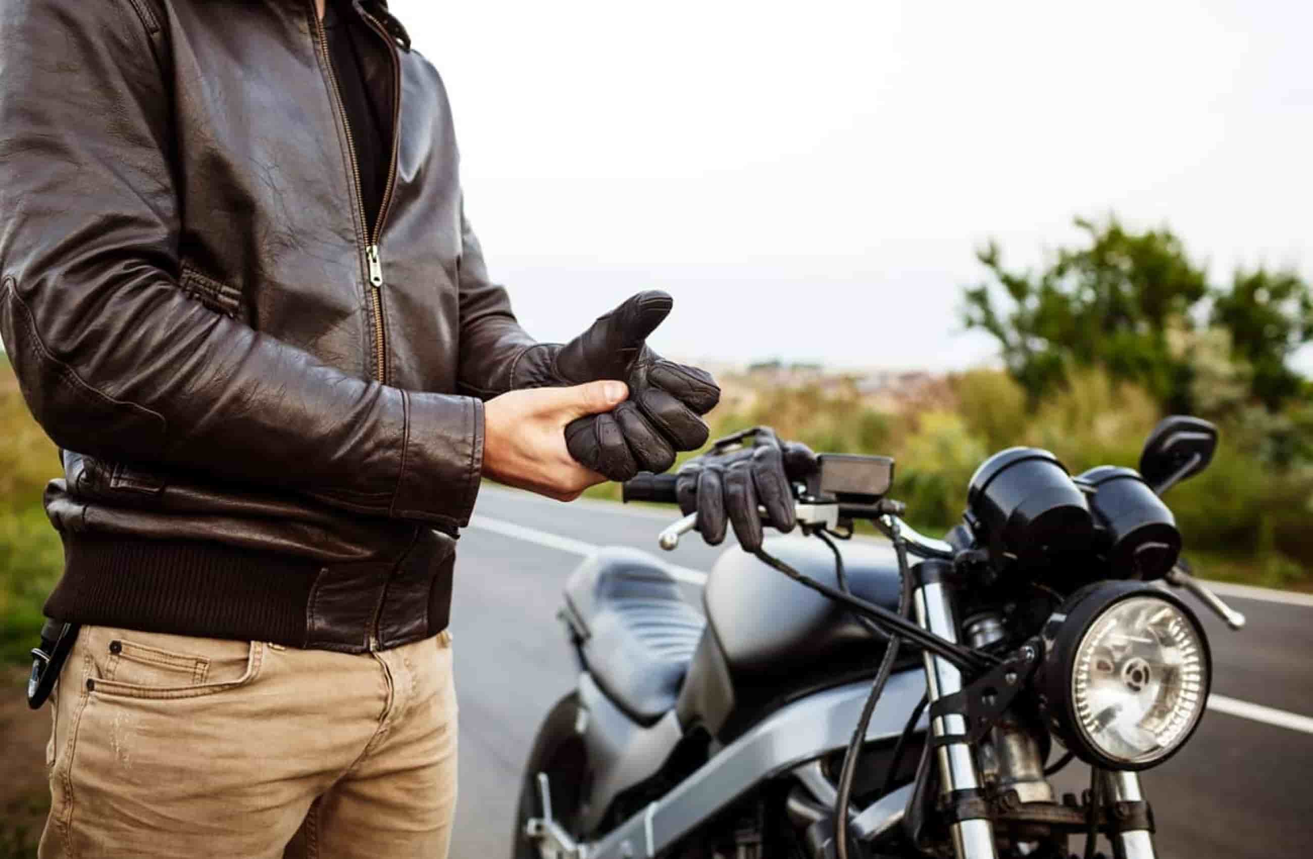 Busto de um homem com jaqueta de couro. Ele está colocando luva de couro em uma das mãos. Ao lado dele tem uma moto parada na estrada.