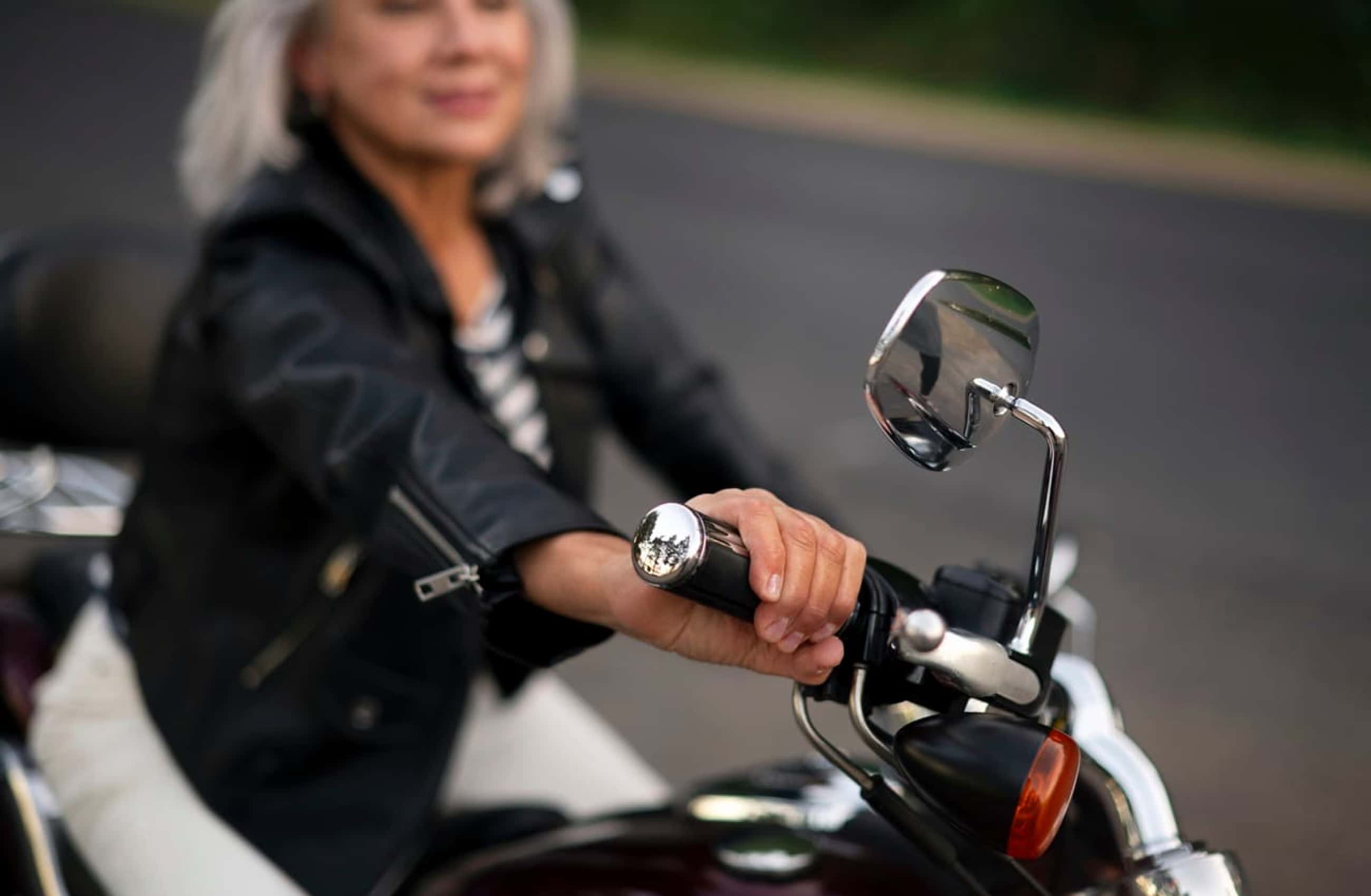 Em primeiro plano a mão de uma mulher está segurando o guidão de uma moto. Ao fundo desfocado, uma mulher banca veste jaqueta de couro enquanto está sentada em uma moto.