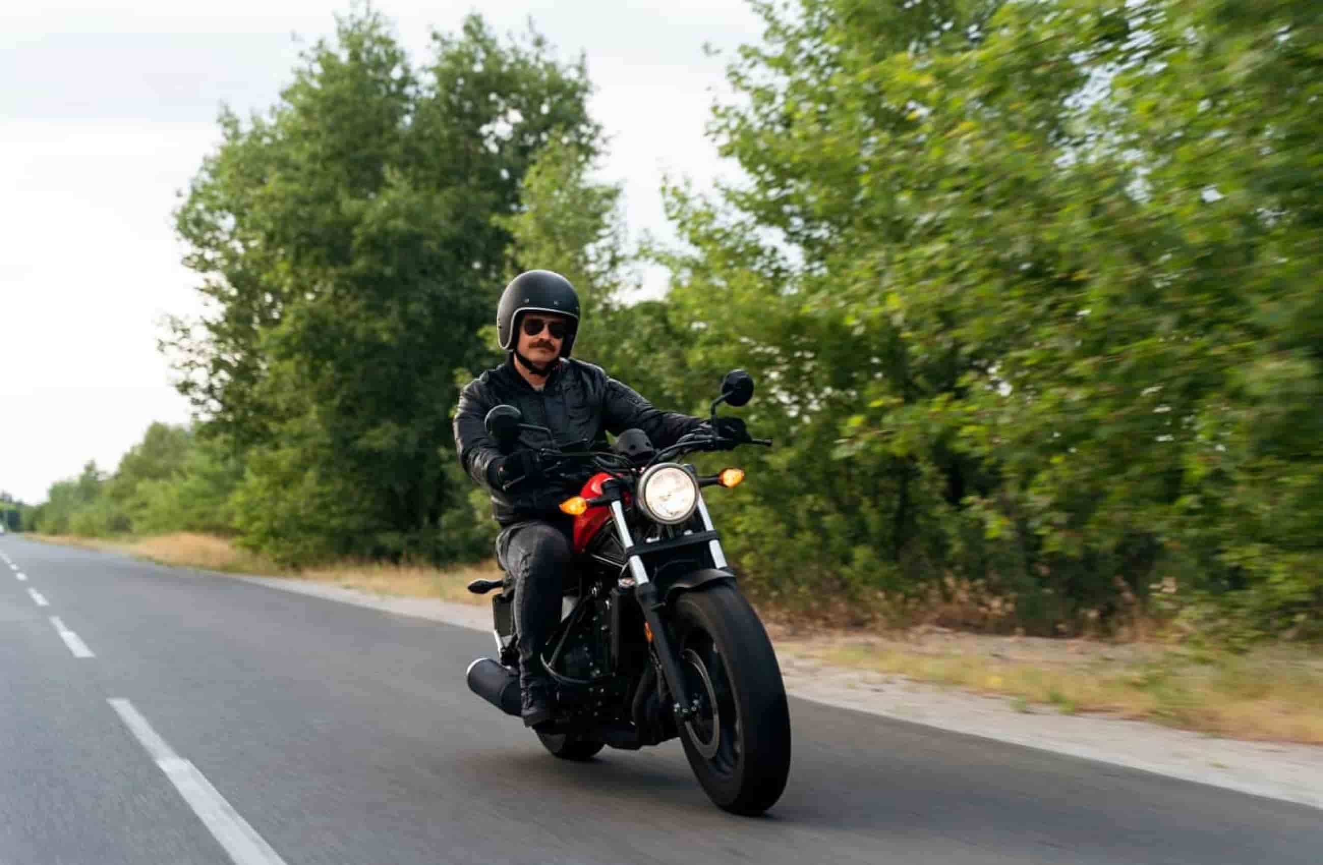 Homem usando óculos escuros, roupa preta e capacete está pilotando uma moto na estrada. Ao fundo, árvores e estrada de asfalto.