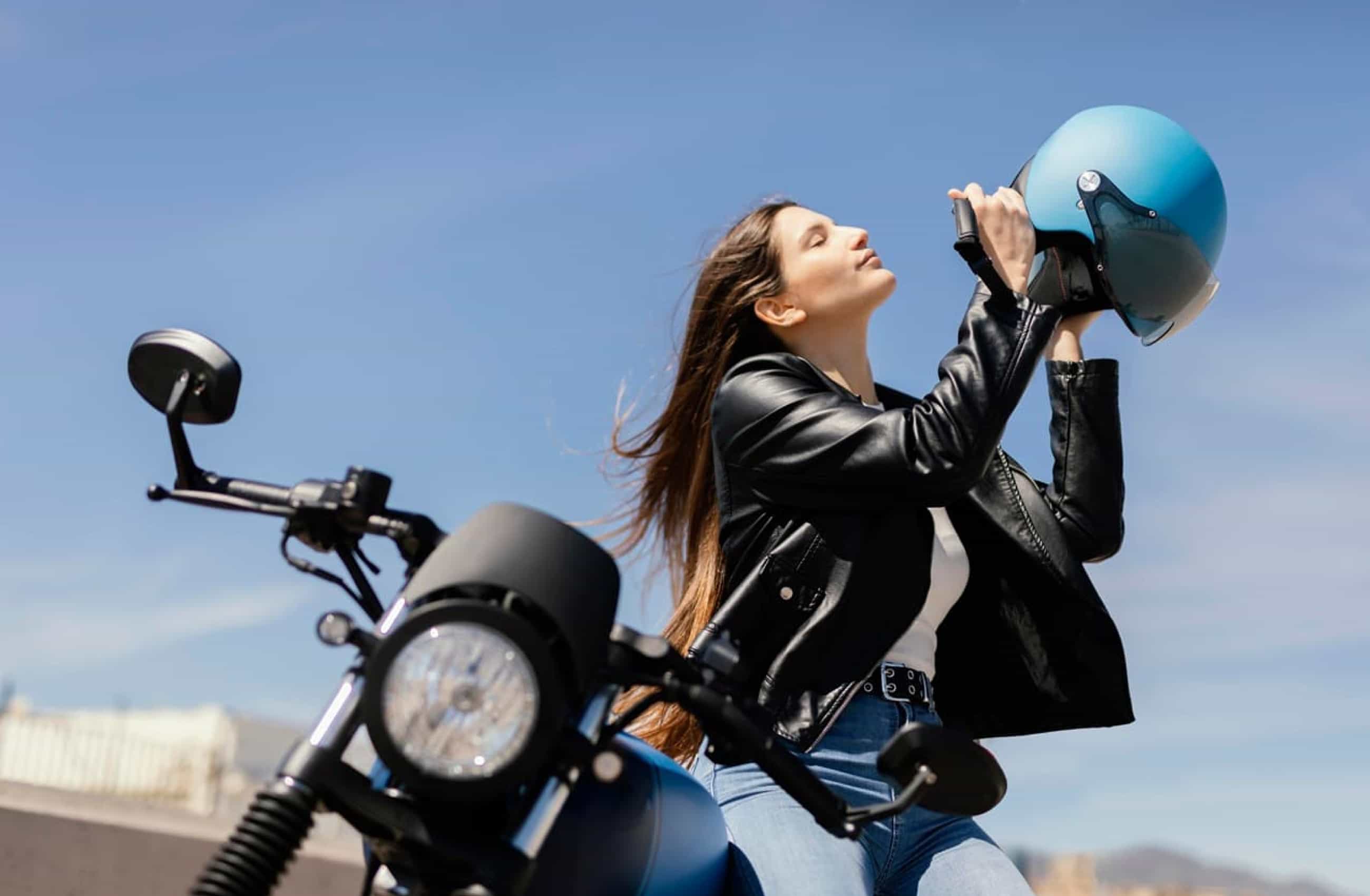 Mulher vestindo uma jaqueta de couro preta com calça jeans e cabelos soltos, está sentada em uma moto. Ela segura um capacete azul para colocá-lo, enquanto está de olhos fechados.