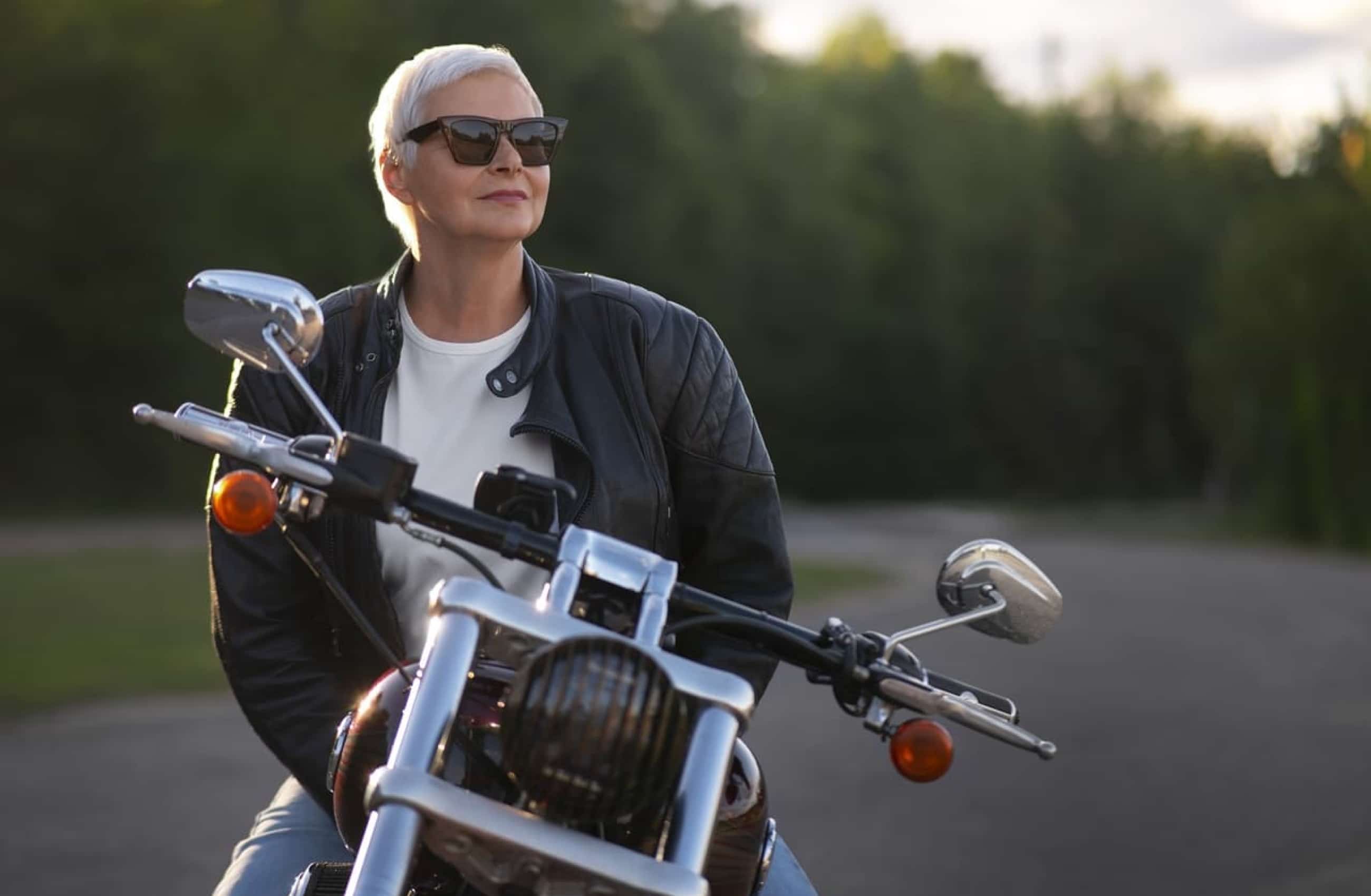Mulher branca com cabelo curto e branco, está usando um óculos de sol e sorri para a paisagem. Ela está encima de uma moto parada.