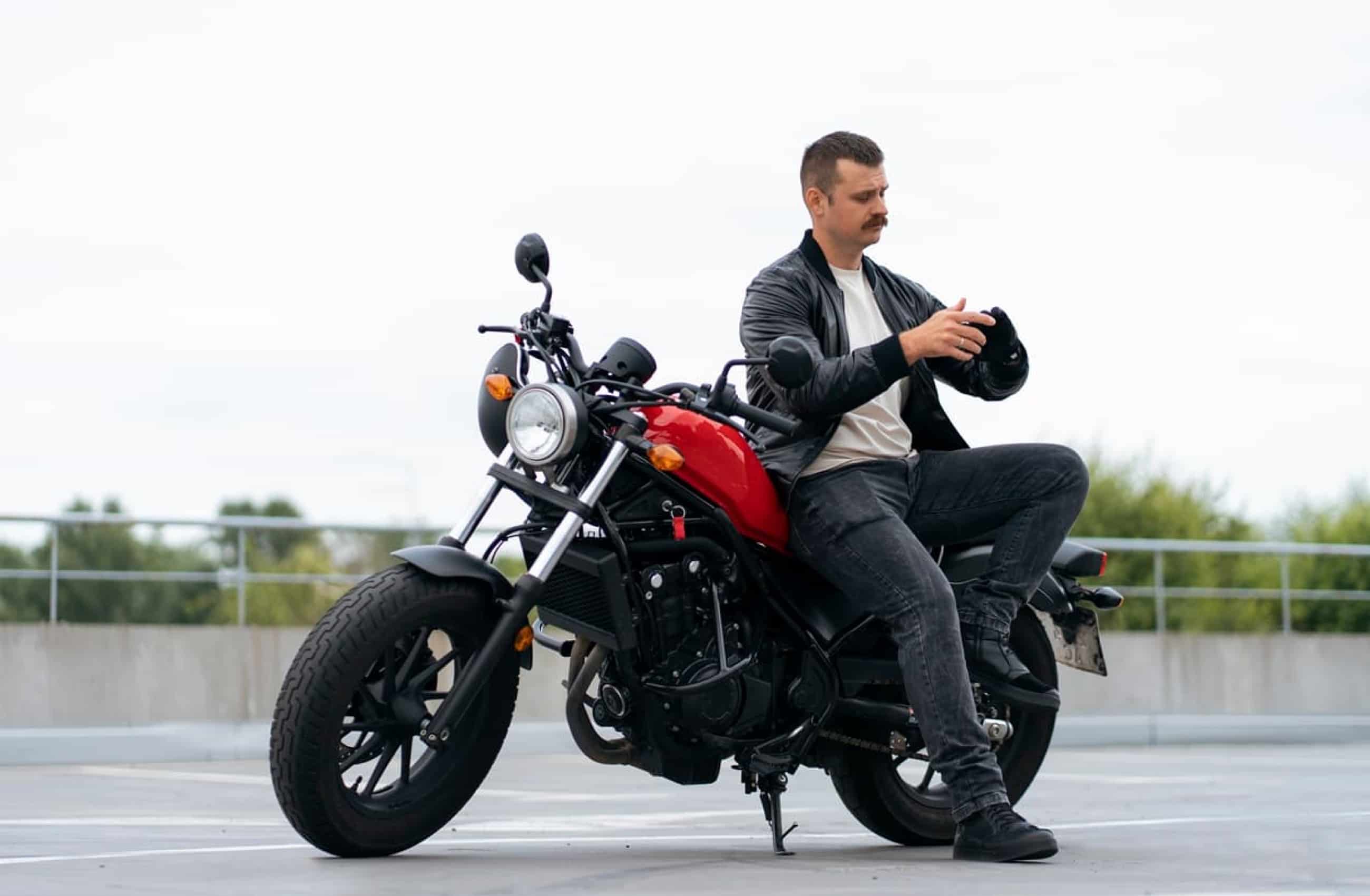 Homem branco usando jaqueta de couro e calça jeans. Ele está colocando uma luva enquanto está sentado parcialmente em uma moto preta e vermelha.