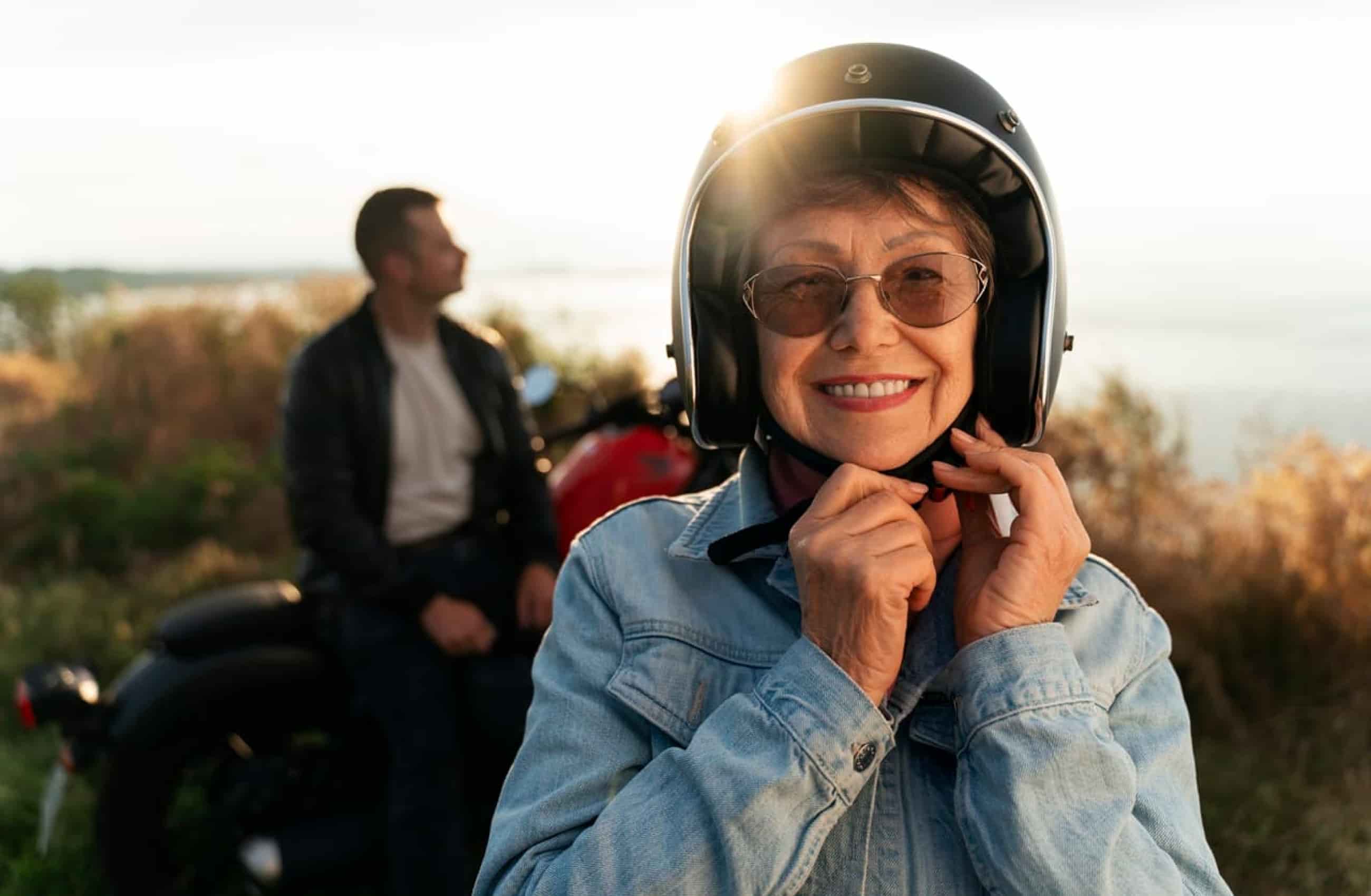 Mulher branca, em torno de 50 a 60 anos de idade, está sorrindo enquanto olha pra câmera, sorri e coloca o capacete de uma moto. Ao fundo, vemos um homem com jaqueta de couro sentado na moto.