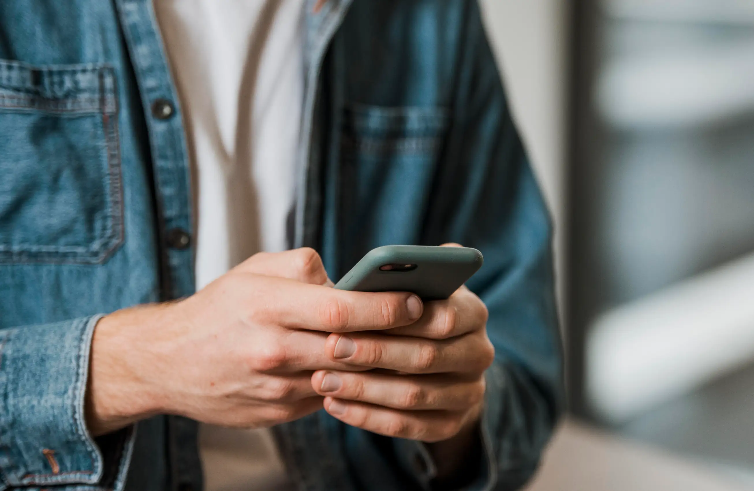 Pessoa de camiseta branca e camisa jeans segurando um smartphone com as duas mãos, concentrada na tela; foco nas mãos e no dispositivo, fundo desfocado.&nbsp;