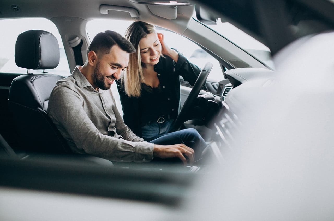 Homem sorridente sentado no banco do motorista de um carro com uma mulher na parte de fora, também sorridente. Eles estão explorando o interior e o console do veículo.