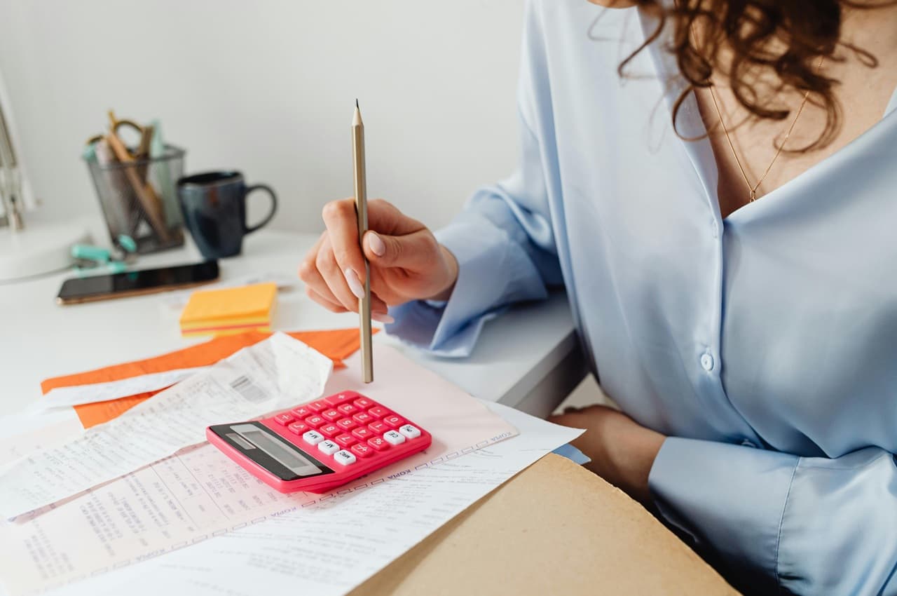 Mulher segurando lápis e usando calculadora rosa, com contas a pagar organizadas sobre a mesa.
