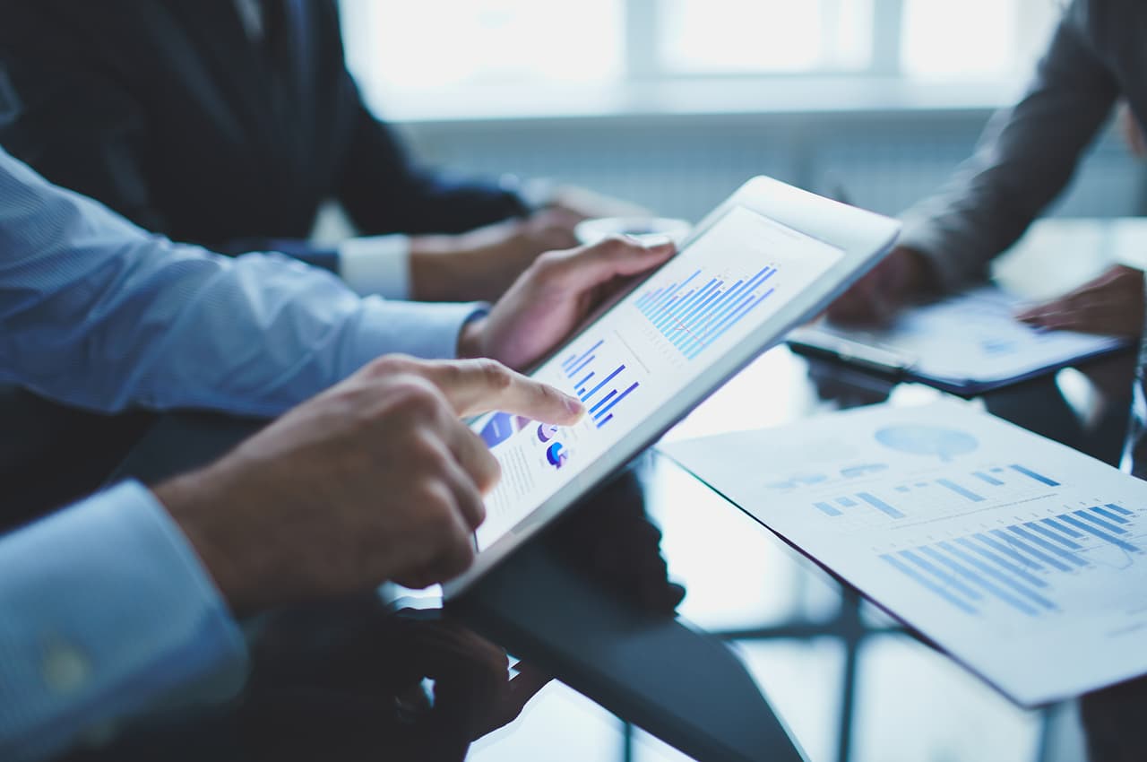 Mãos de executivos analisando gráficos e relatórios em um tablet durante reunião sobre contrato de royalties.&nbsp;