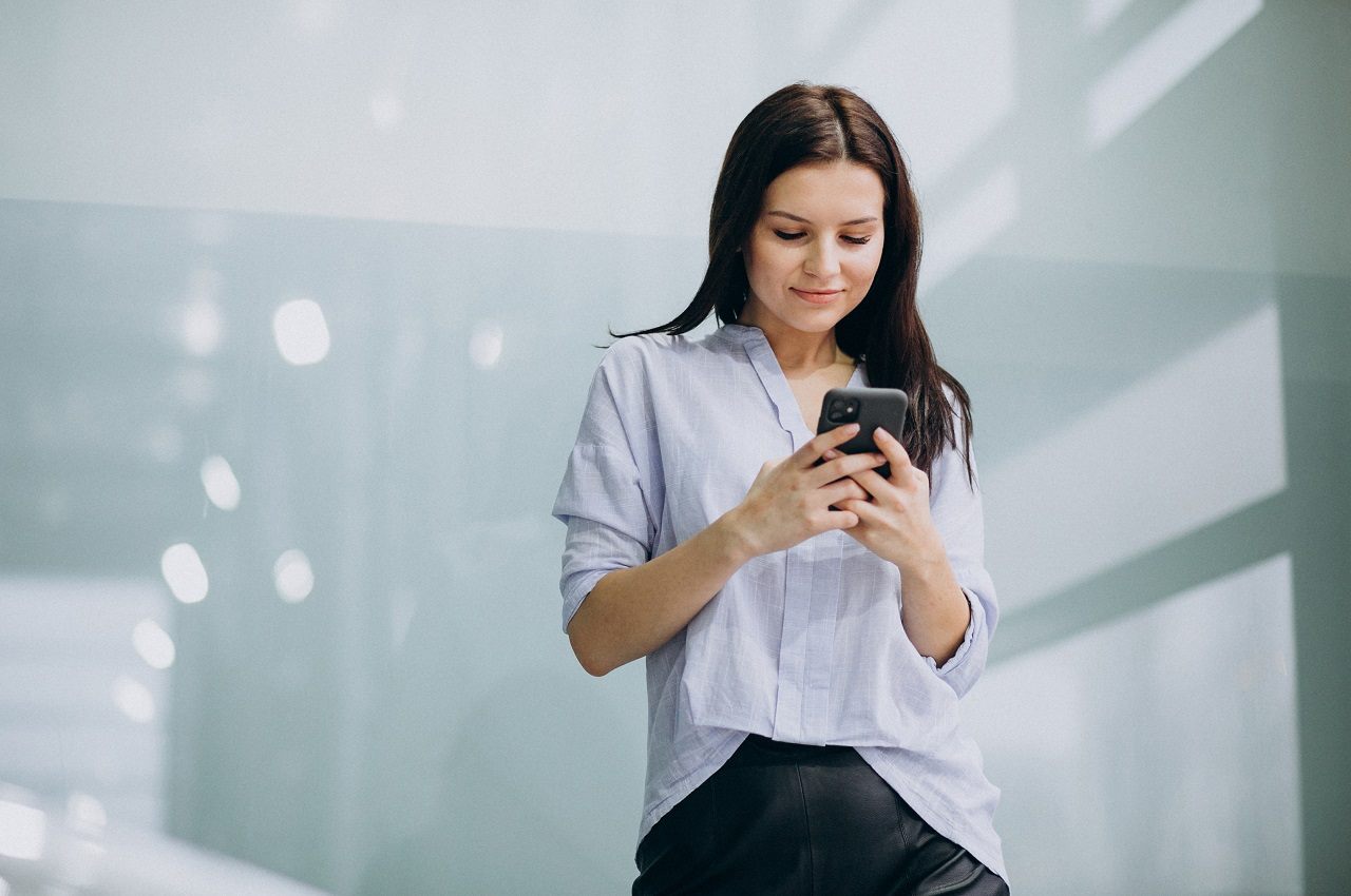 Mulher sorridente, em pé vestindo calça social preta e camisa social branca. Ela utiliza um celular.