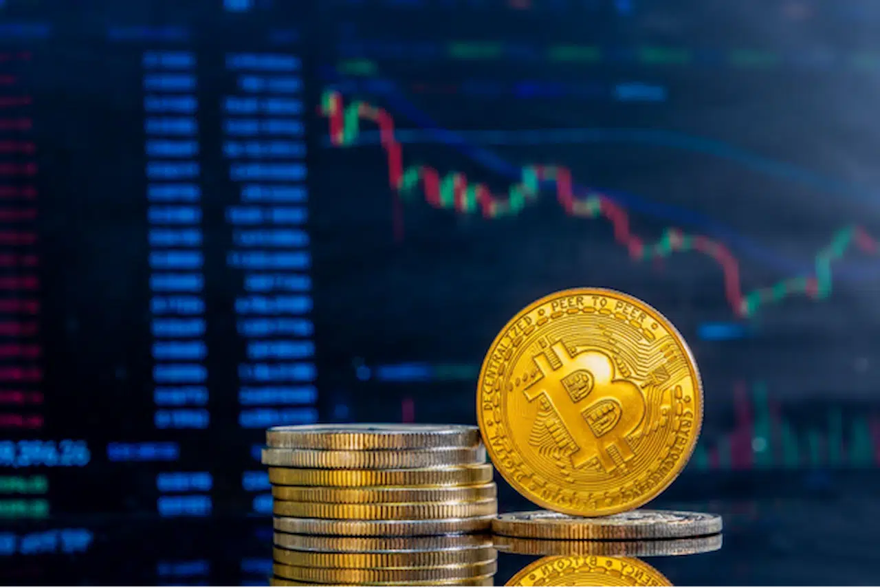 Pilha de moedas douradas, com uma em pé virada para a frente com um símbolo do bitcoin. Ao fundo, está uma tela com gráficos financeiros.