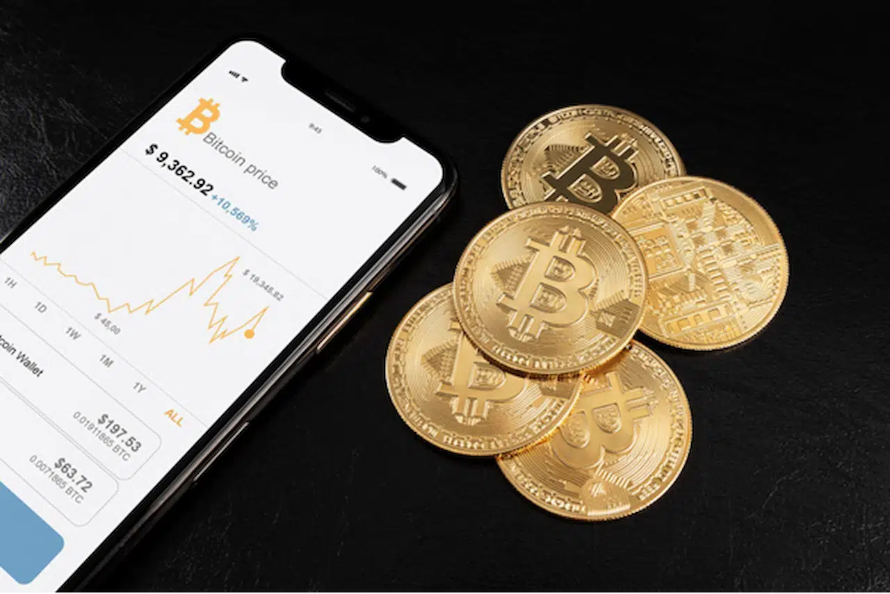 Celular mostrando a tela com o saldo de uma conta de bitcoin, com gráfico e valores financeiros. Ao lado, estão moedas douradas representando as criptomoedas.&nbsp;