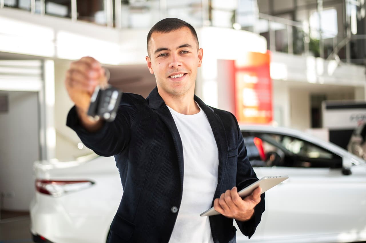 Homem sorridente de blazer segurando um tablet e estendendo a chave de um carro em uma concessionária.