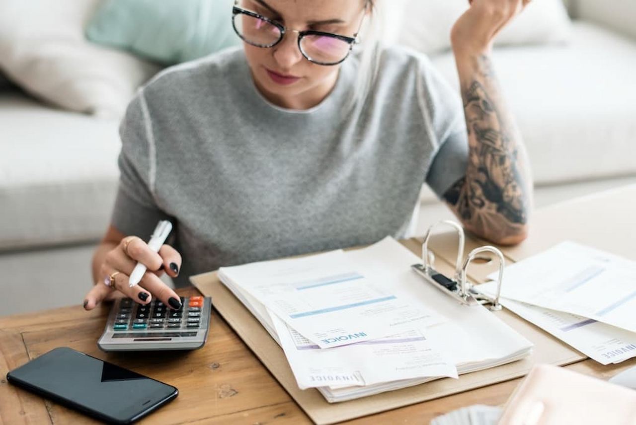 Mulher com expressão concentrada, óculos de grau e calculadora conferindo o que é IPCA. A mulher tem tatuagens no braço esquerdo e vários papéis em um fichário em cima da mesa.