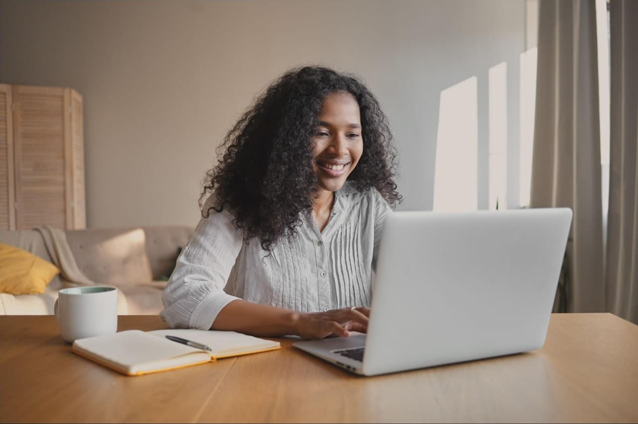 Mulher sentada à uma mesa, utilizando um notebook, com um caderno aberto ao seu lado e uma xícara.
