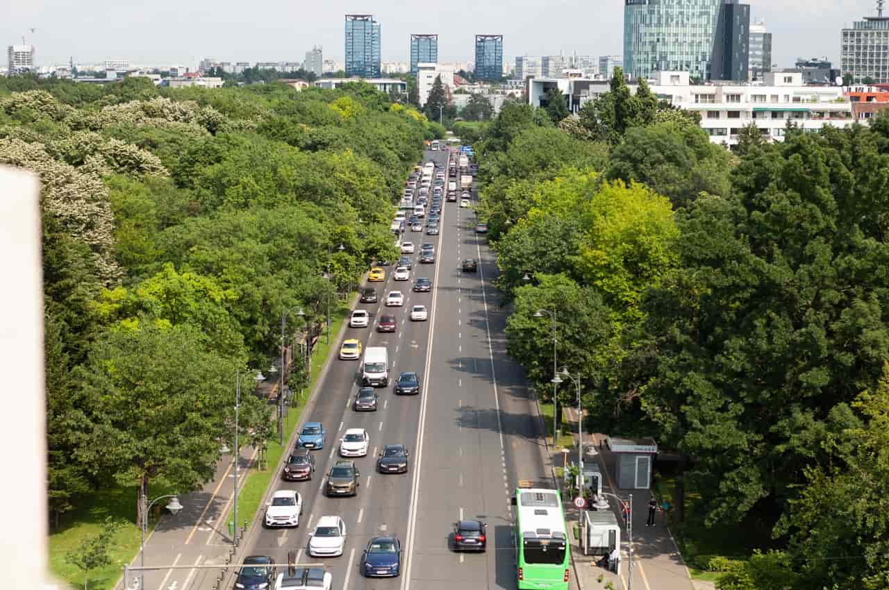 Vista aérea de uma avenida movimentada com carros e ônibus, cercada por densa vegetação, com o horizonte de uma cidade ao fundo.