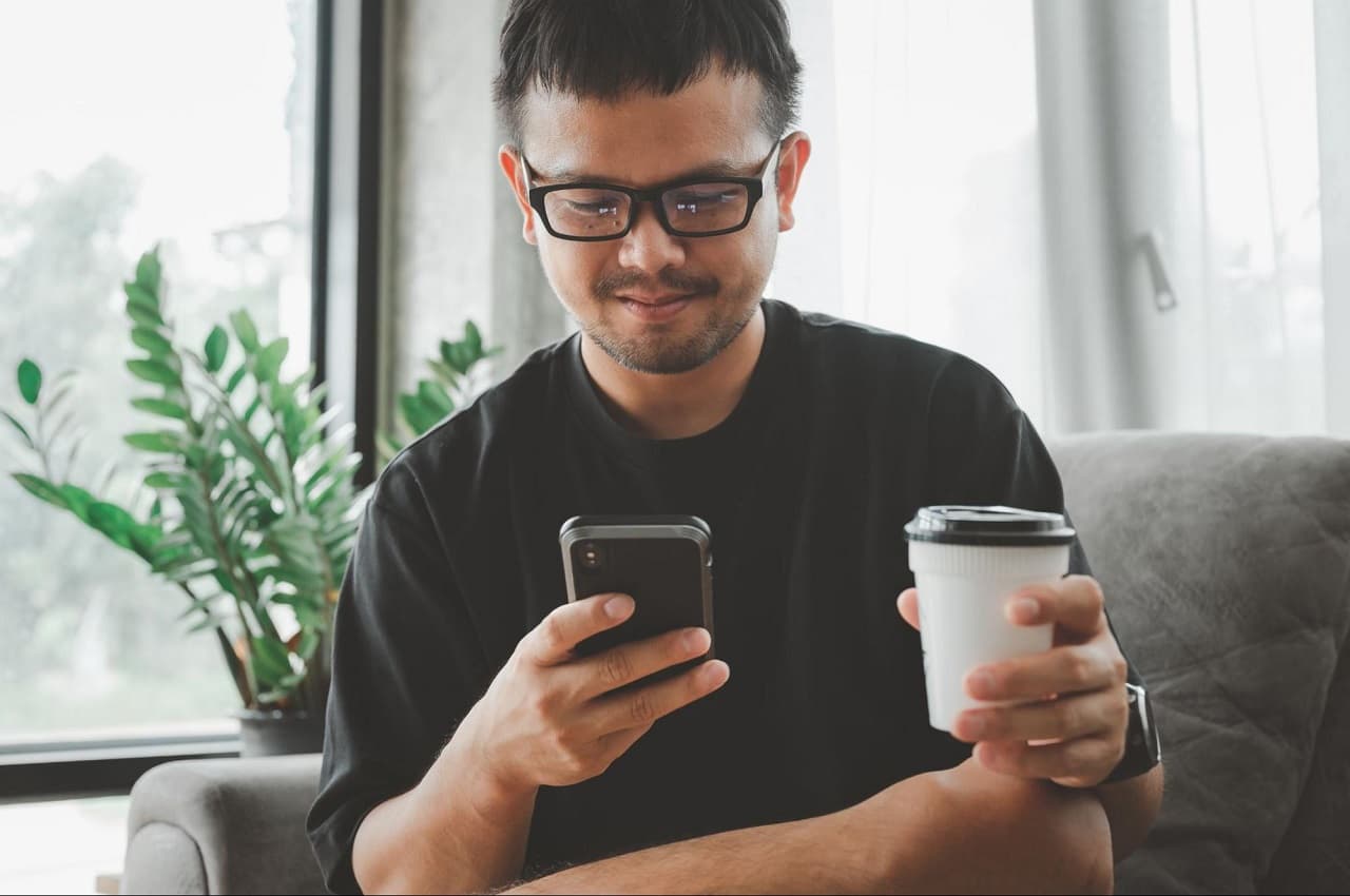 Homem sorridente, vestindo blusa básica preta e copo térmico de café, acessando saque-aniversário pelo celular. Ele está sentado no sofá de sua casa. 