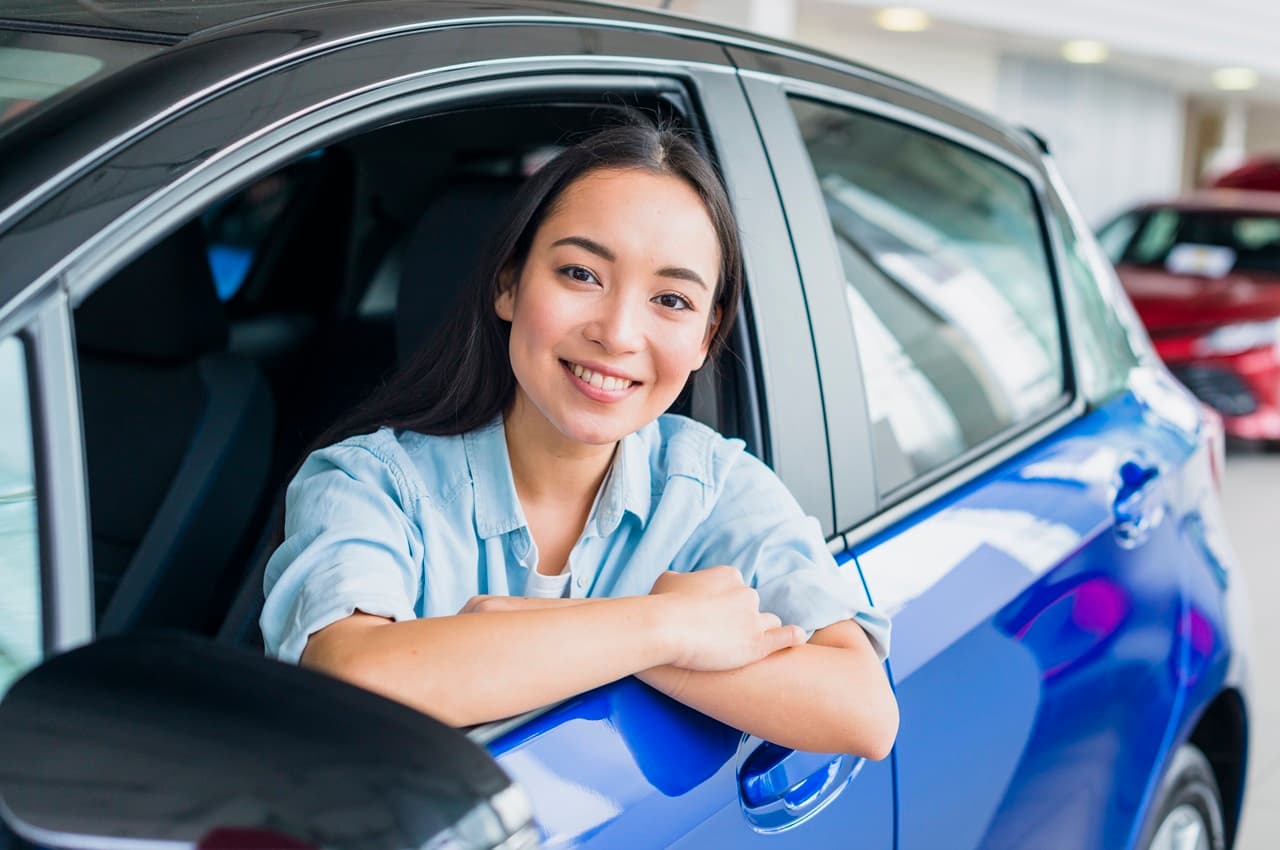 Mulher jovem sorrindo enquanto está debruçada na janela do motorista de um carro azul, que se encontra no interior de uma concessionária.