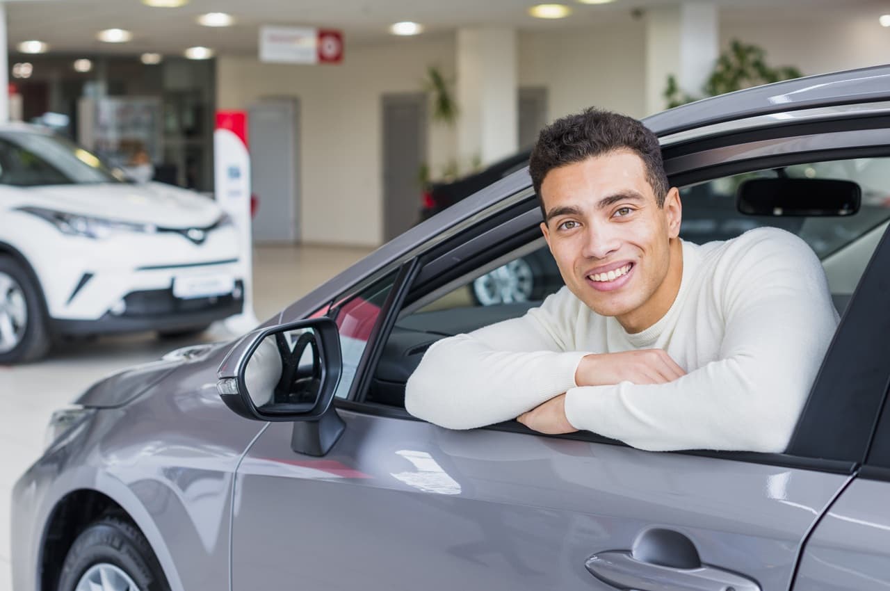 Homem jovem sorridente com os braços apoiados na janela aberta de um carro cinza em uma concessionária.