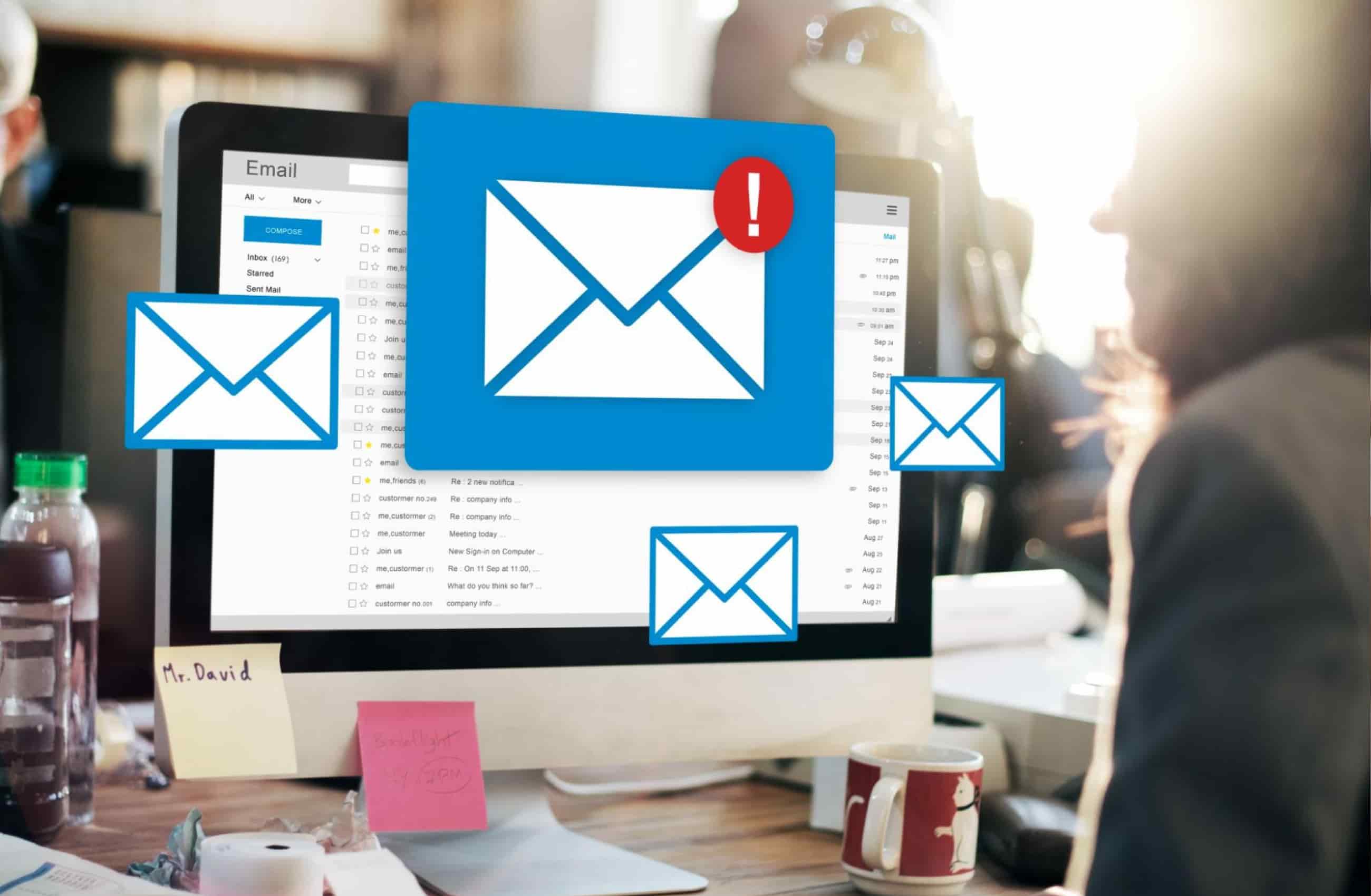 Pessoa trabalha em um computador com a caixa de e-mail aberta, cercada por ícones de envelope — um deles com alerta vermelho, simbolizando mensagens suspeitas e o risco de phishing no ambiente corporativo.