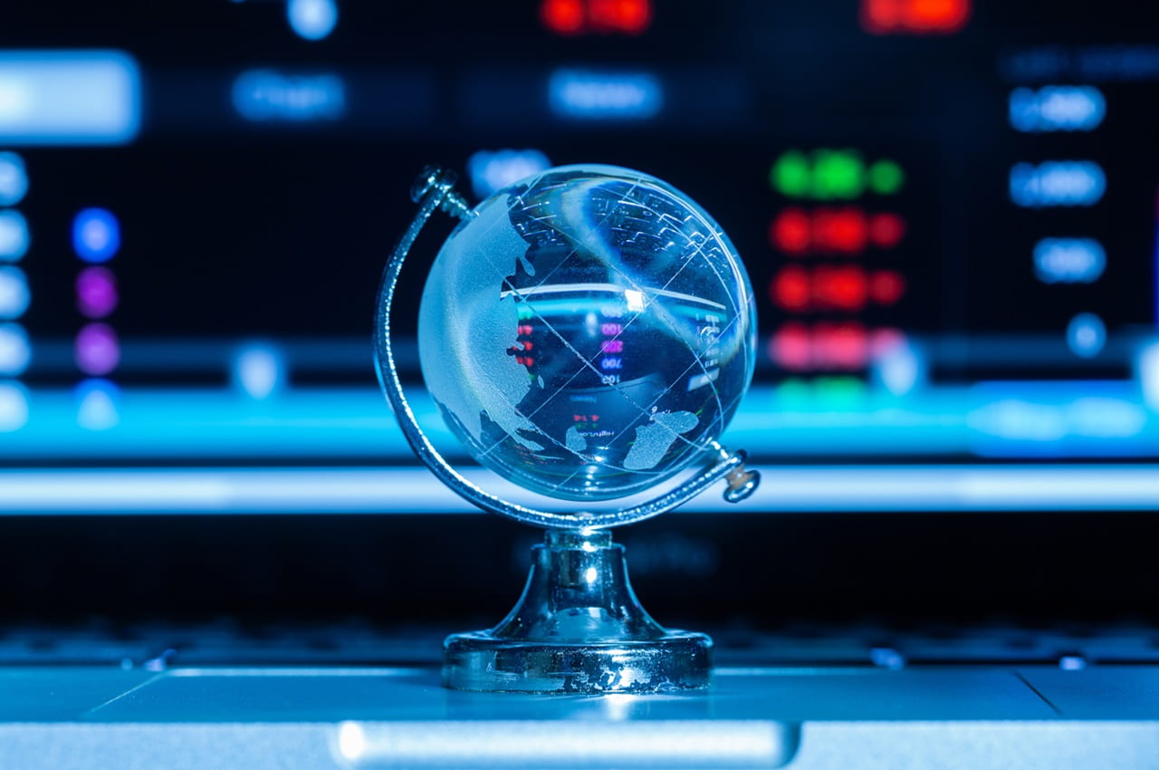 Globo terrestre de vidro, apoiado em suporte metálico, com um fundo de tela com gráficos financeiros coloridos, que refletem no globo e simbolizam operações onshore e offshore.