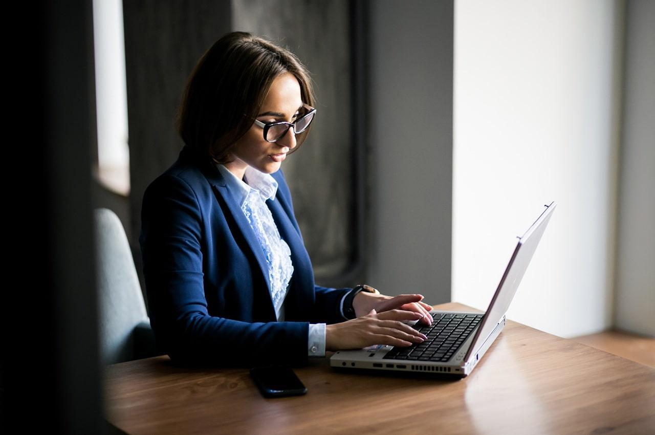 Mulher de óculos e cabelos castanhos, vestindo blazer azul, enquanto está sentada, trabalhando em um notebook e pesquisando sobre operações compromissadas.