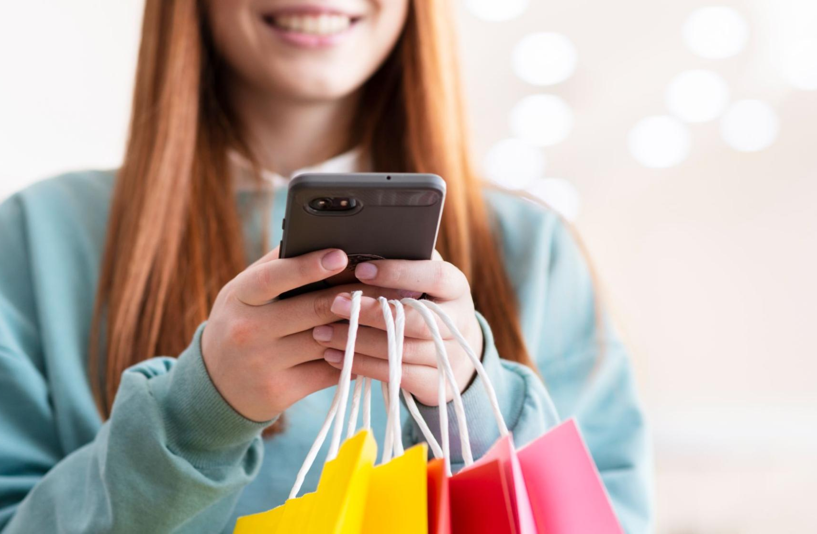 Mulher de cabelos ruivos, segurando várias sacolas de compras coloridas em uma das mãos, enquanto usa um smartphone com a outra. 
