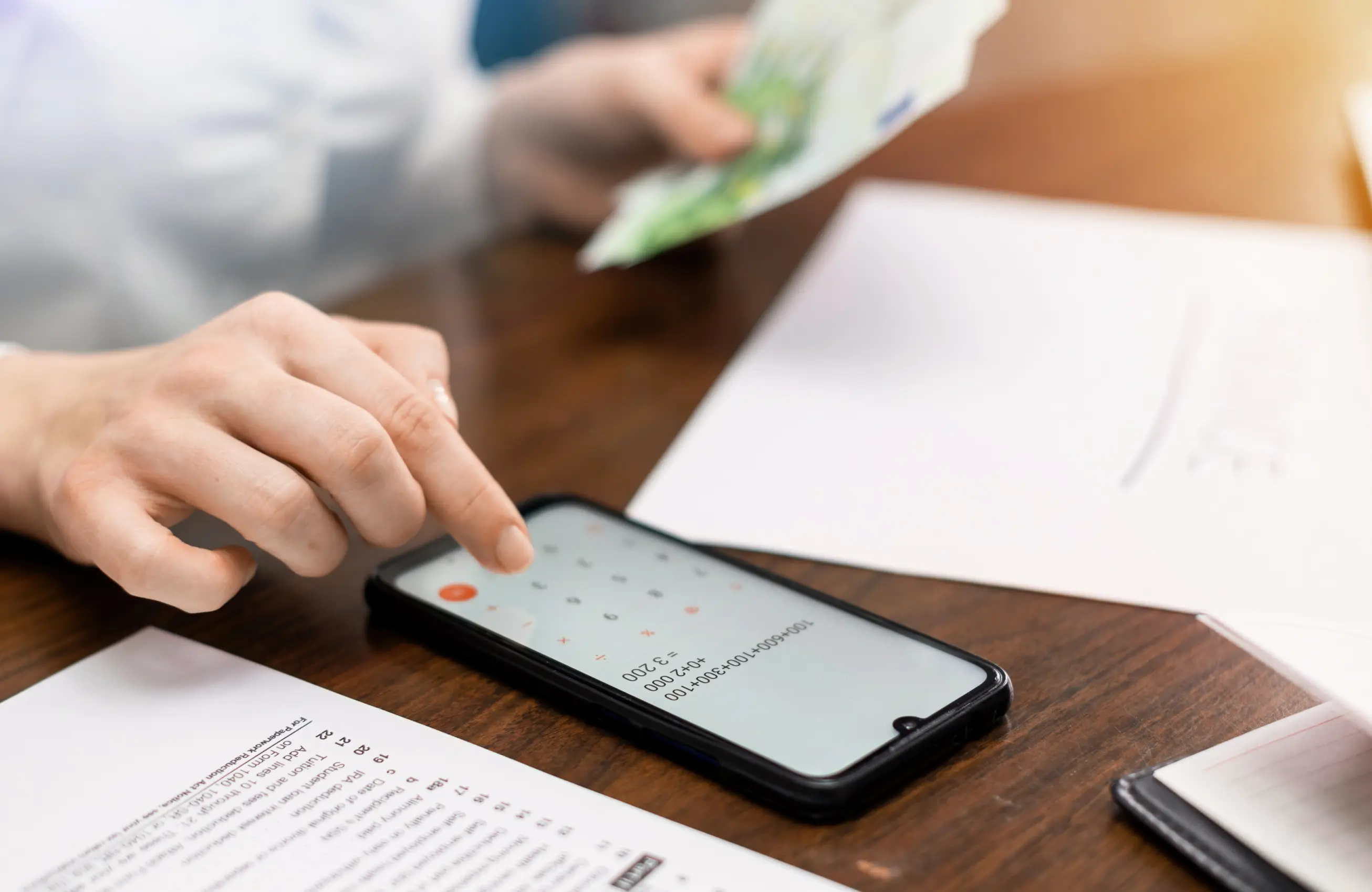 Pessoa usando um smartphone na mesa de trabalho, com papéis e caderno ao redor; outra pessoa ao fundo segurando dinheiro.