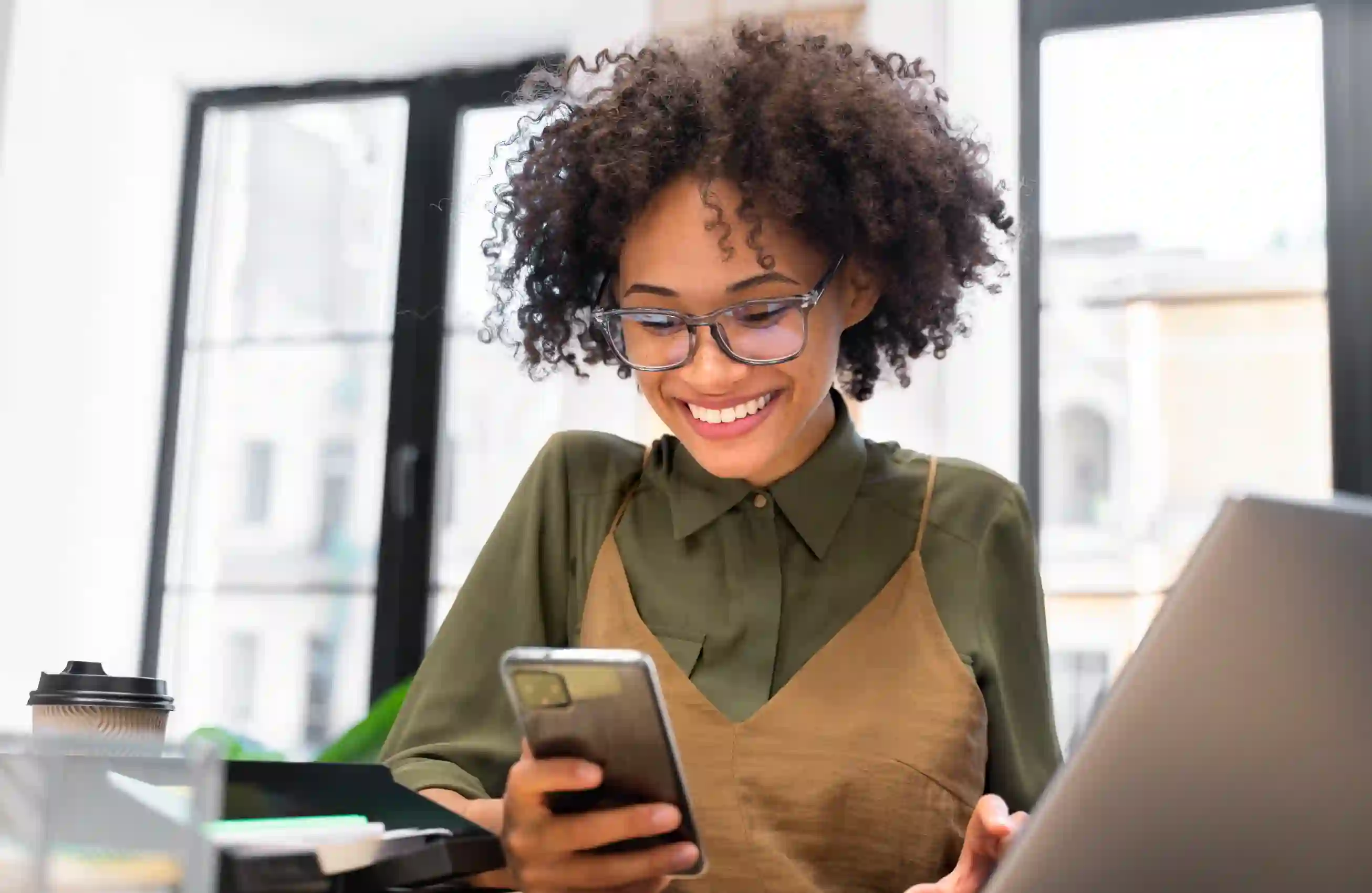 Mulher jovem com cabelos cacheados e óculos, sorrindo, está olhando para o celular enquanto trabalha em uma mesa com laptop aberto e itens de escritório ao redor.