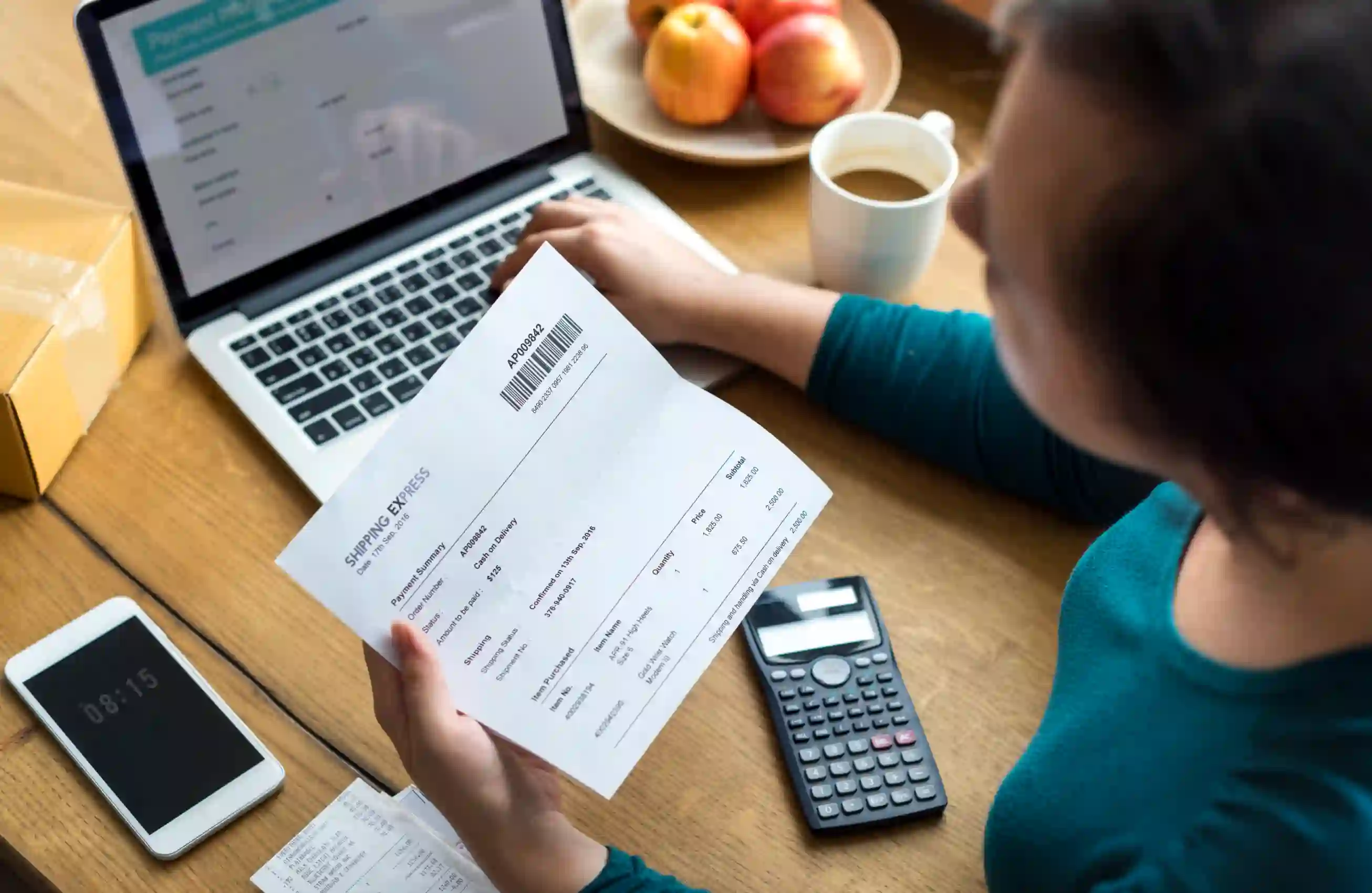 Pessoa segurando uma fatura com código de barras, em frente a um laptop aberto, com smartphone, calculadora, café e maçãs ao fundo sobre a mesa de madeira.&nbsp;