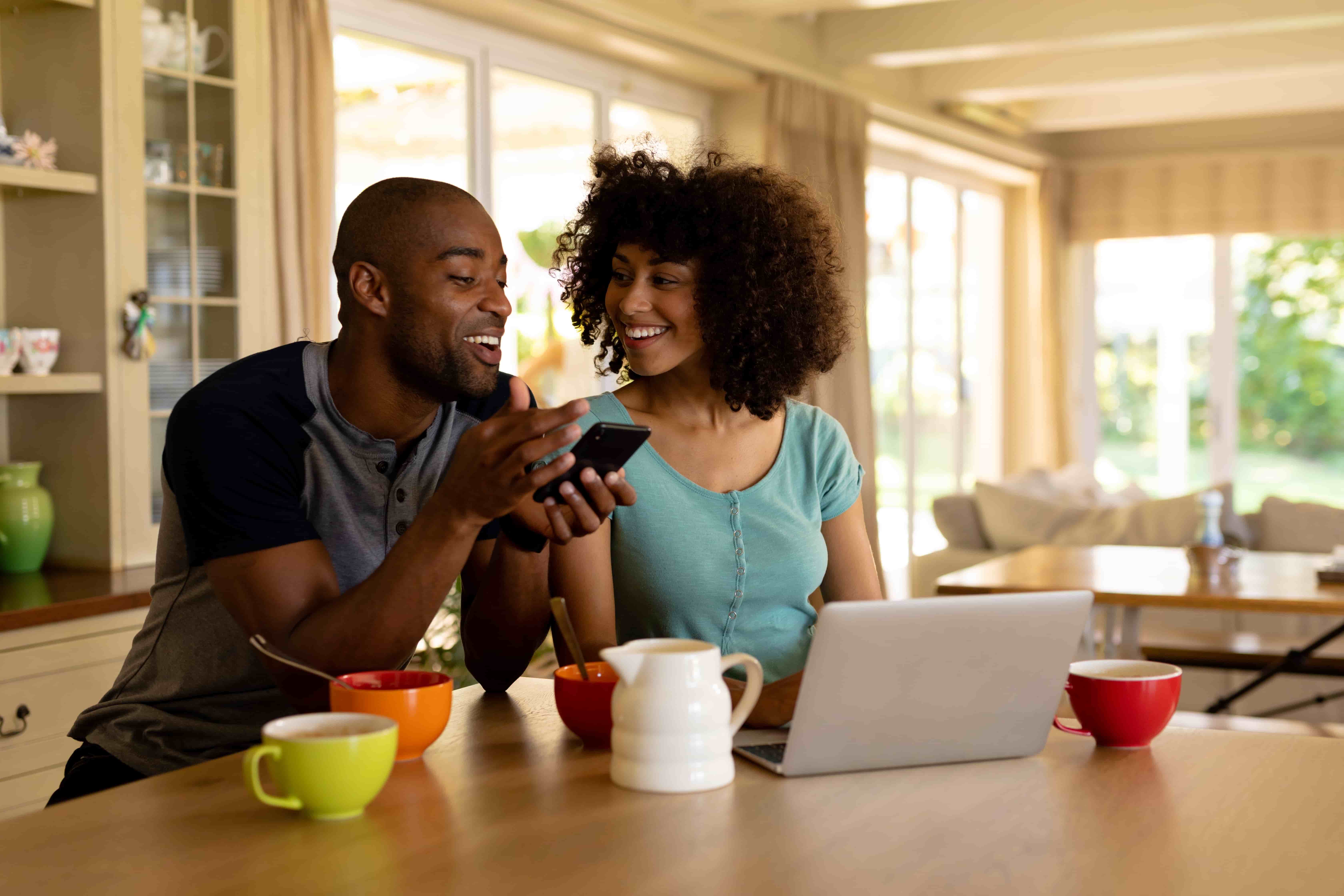 casal na bancada de uma cozinha sorrindo, o homem está segurando um celular nas mãos e mostrando a tela para a mulher