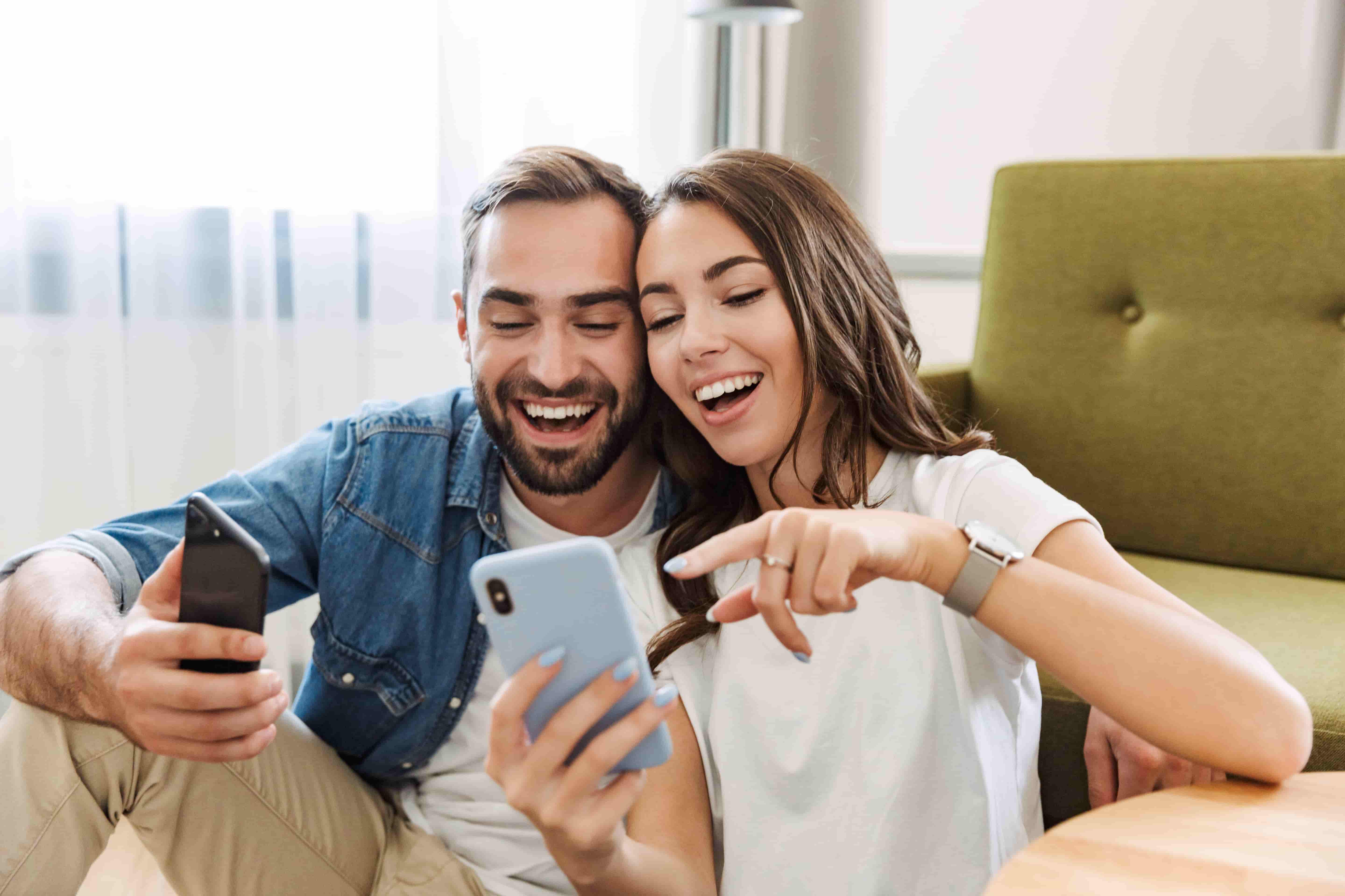 Homem e mulher sentados no chão, um ao lado do outro. Mulher está segurando o celular e apontando para tela, mostrando para o homem.