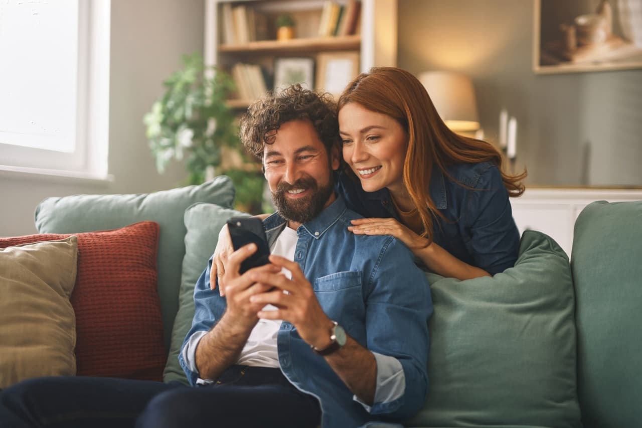 homem e mulher sentados no sofá sorrindo, olhando o celular.
