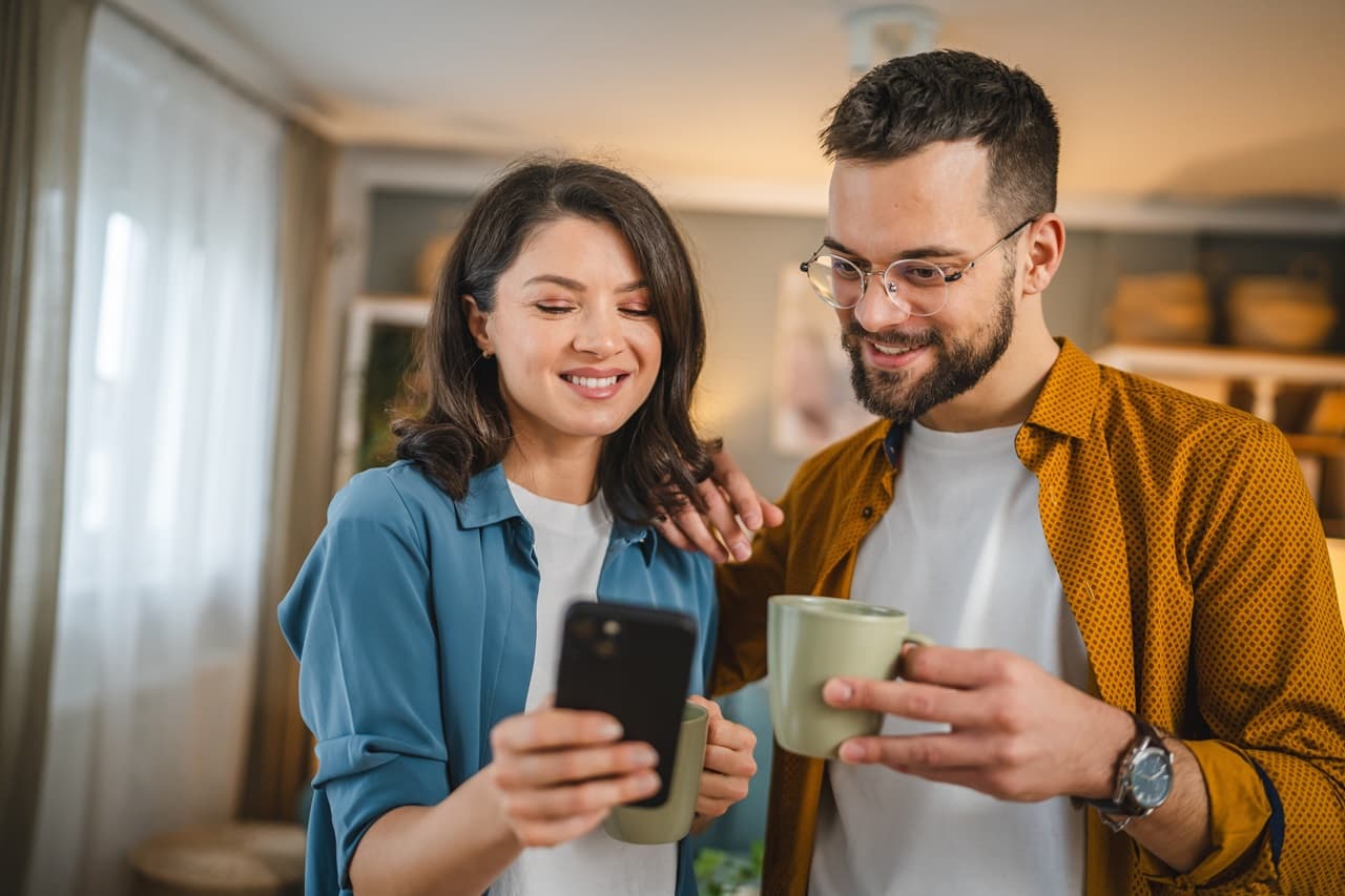 mulher e homem em pé lado a lado, a mulher está segurando um celular na mão e o homem está segurando uma xícara.