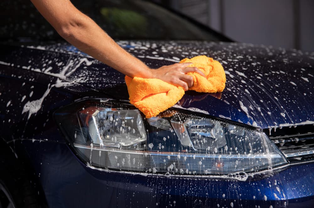Mão passando pano laranja na lataria de um carro azul ensaboado.