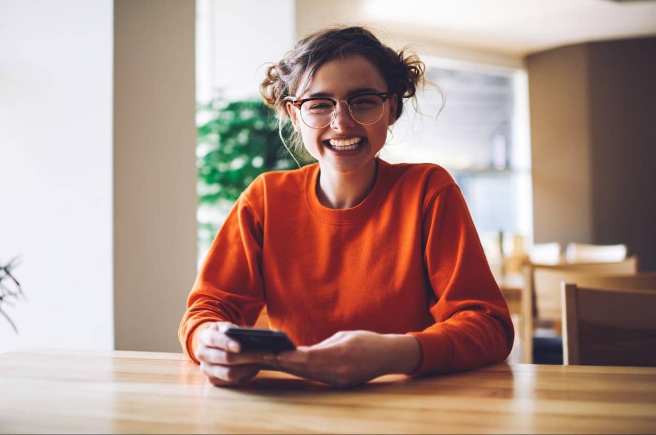 Mulher sorridente vestindo moletom laranja e coques no cabelo, enquanto acessa empréstimo FGTS pelo app no celular.