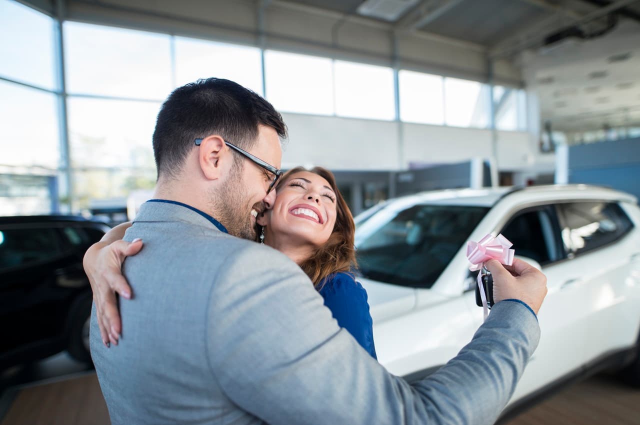 Homem abraçando uma mulher sorridente, enquanto segura uma chave de carro com laço rosa, em uma concessionária.