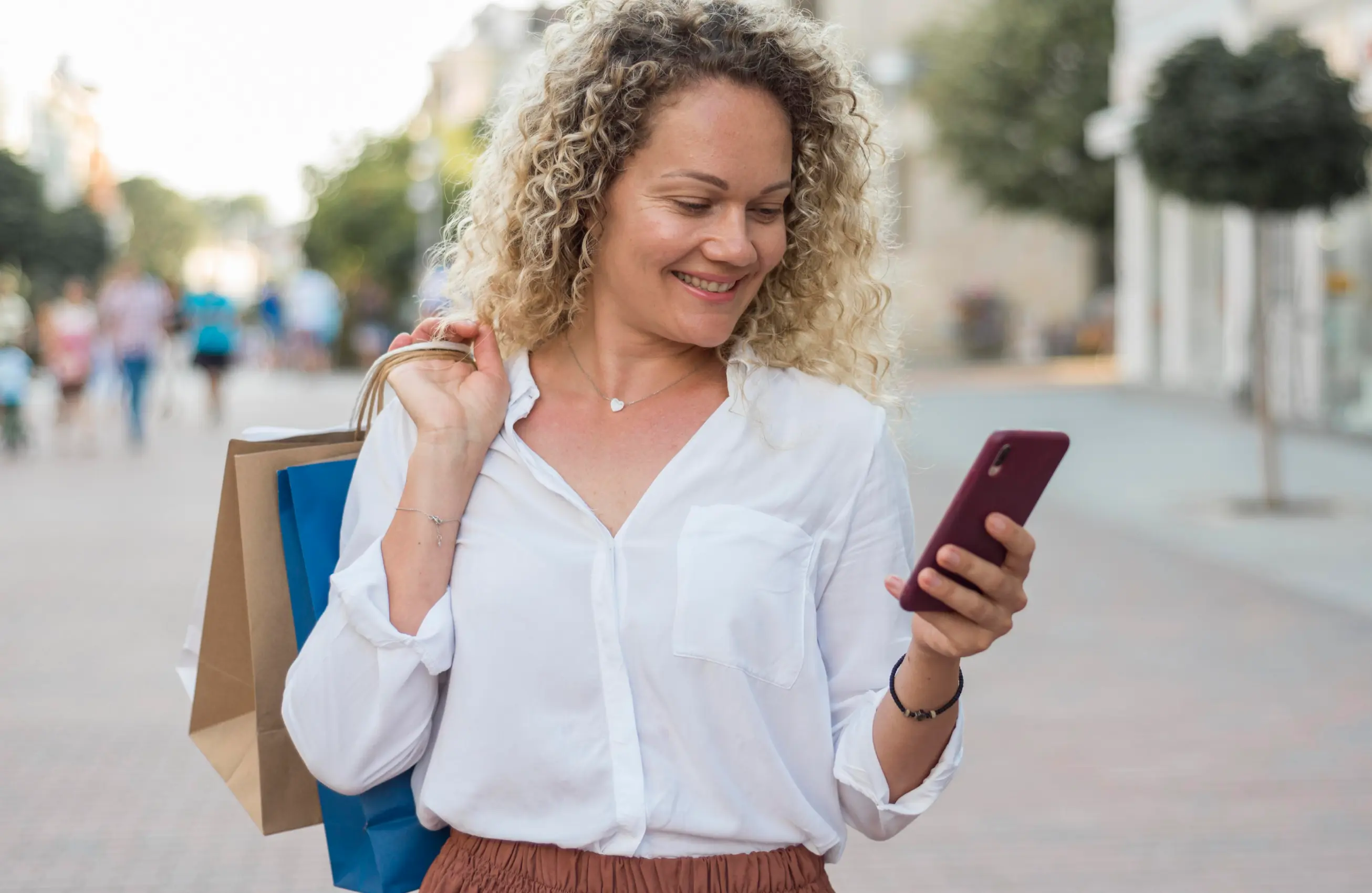 Mulher carregando sacolas de papel sobre o ombro com uma mão, e com a outra segurando um celular, olhando para ele.