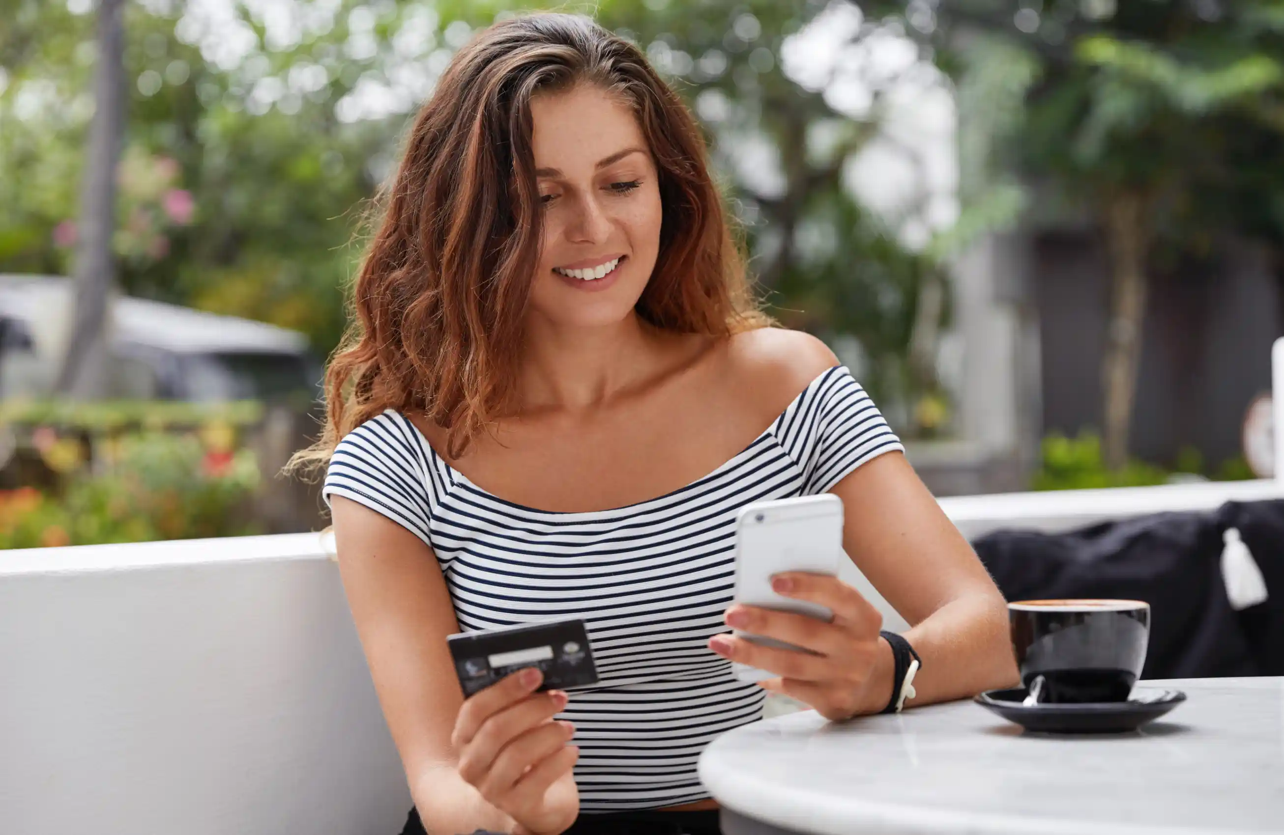 Mulher sentada em uma escada segurando em uma mão um cartão e em outra um celular.&nbsp;