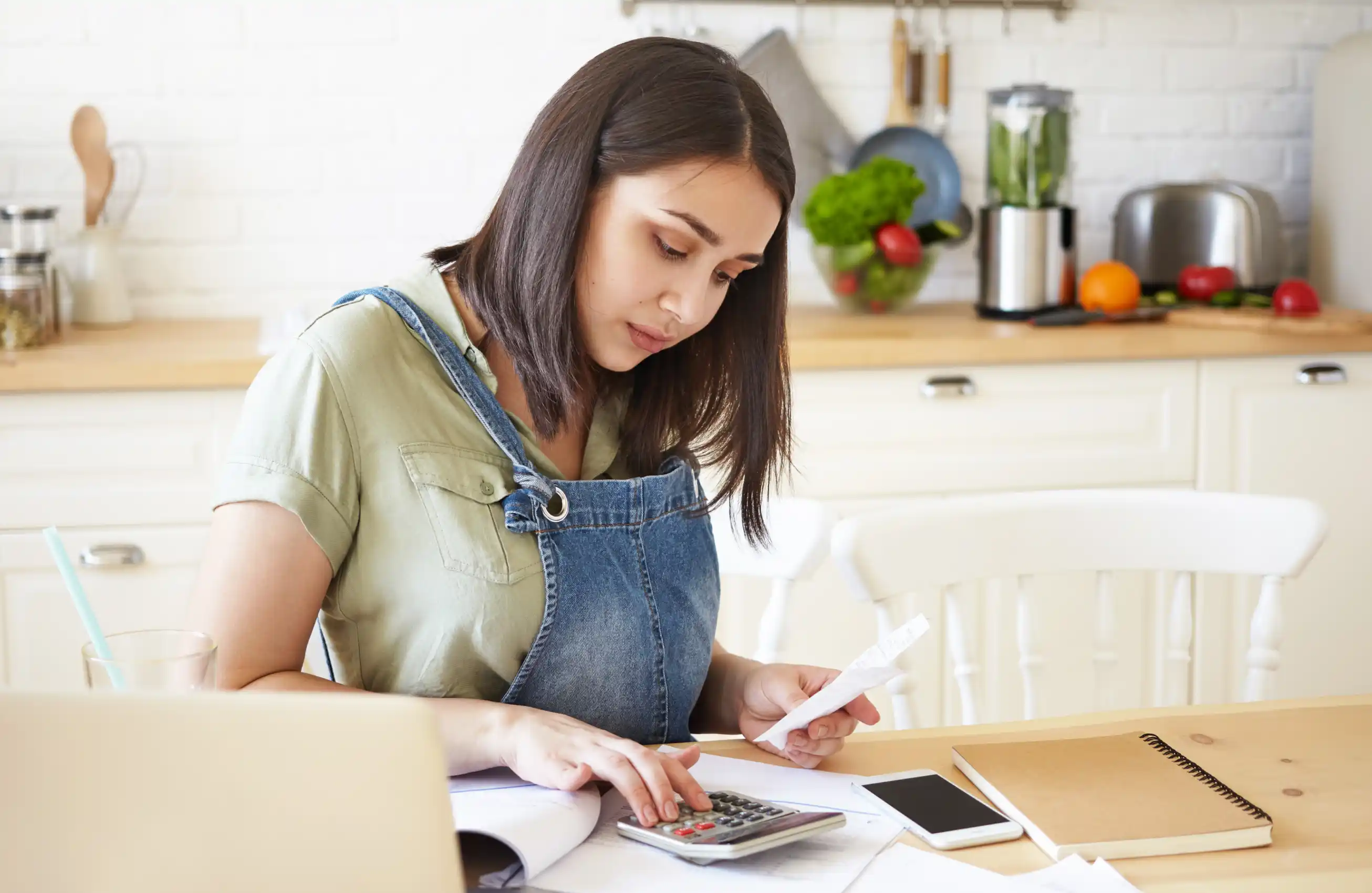 Uma imagem de uma mulher sentada em uma mesa de madeira na cozinha enquanto faz contas em uma calculadora.&nbsp;
