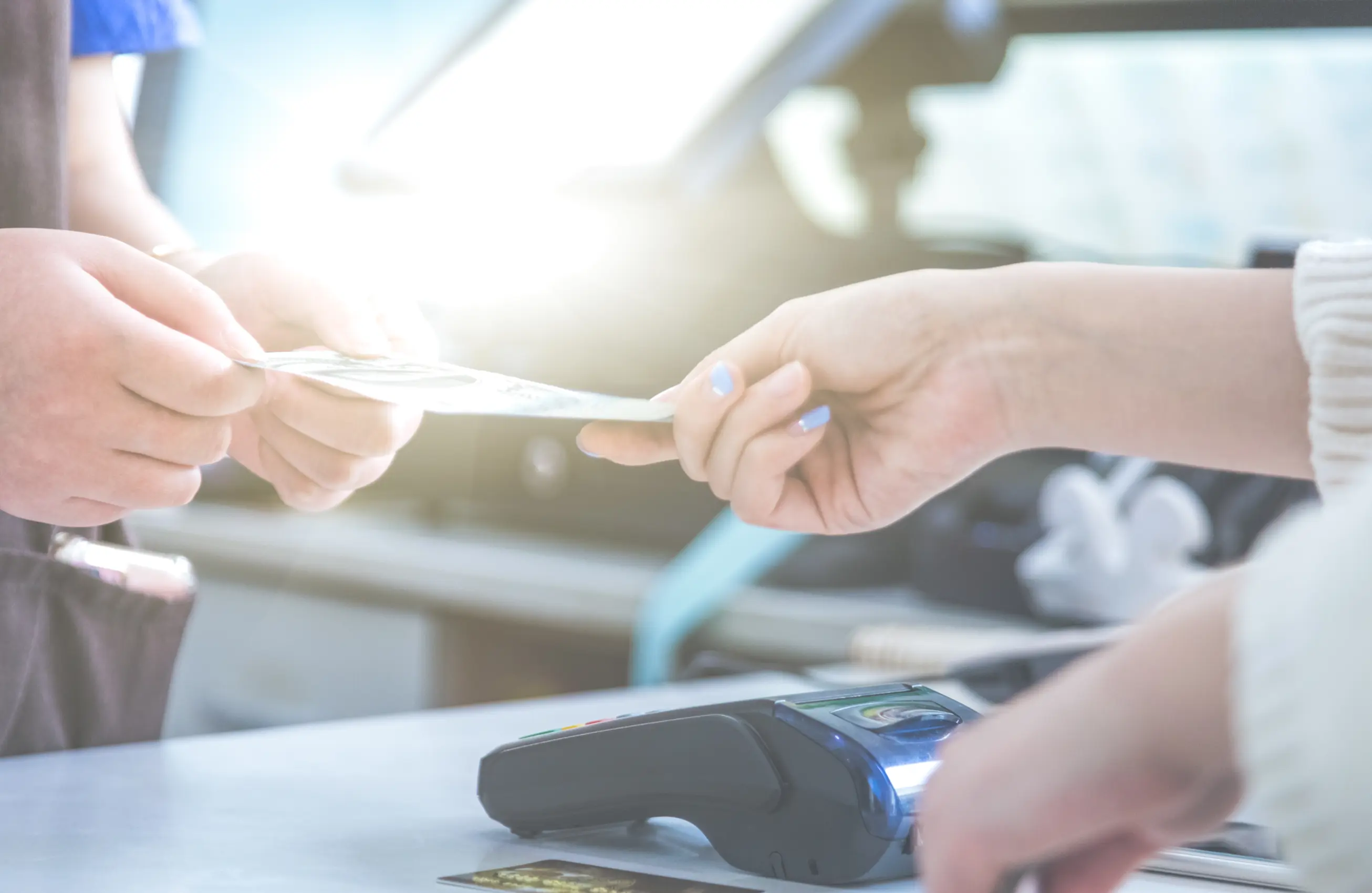 Duas mãos segurando uma nota sendo dada por outra pessoa, sobre uma bancada com uma máquina de cartão.