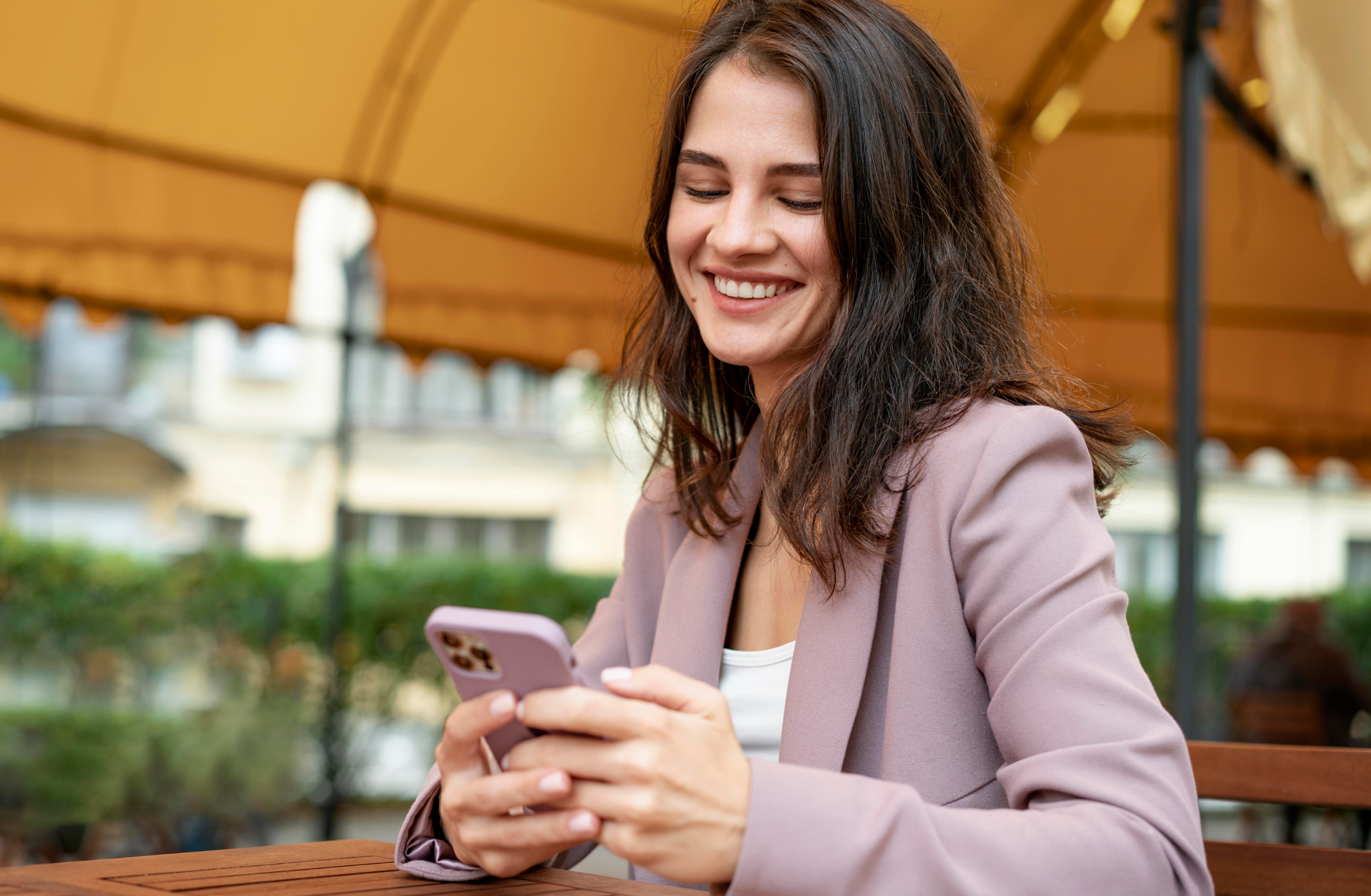 Uma mulher jovem e sorridente, vestindo um blazer lilás, manuseia um smartphone com as duas mãos enquanto está sentada em uma mesa de madeira ao ar livre. 