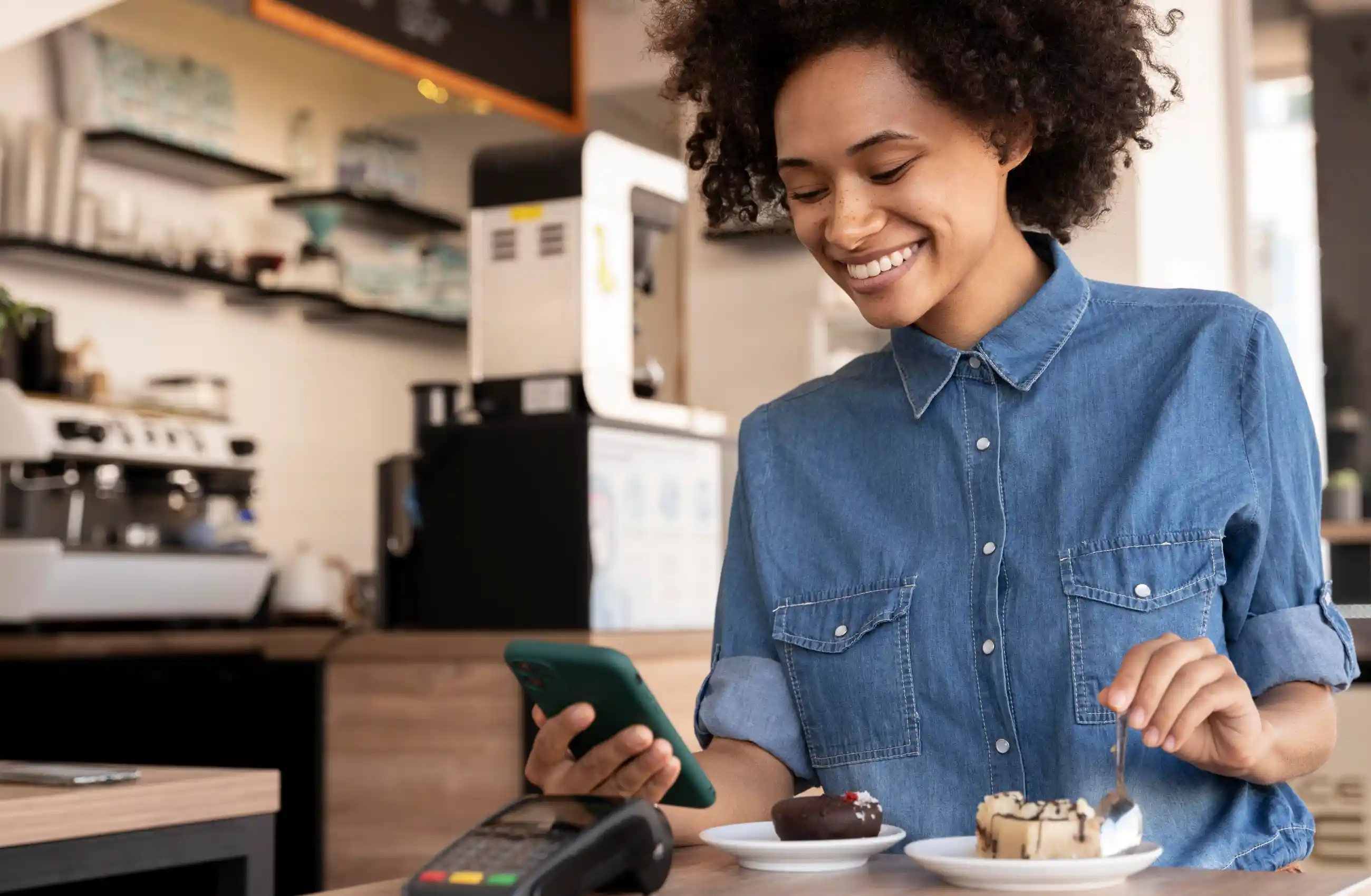 Mulher sorridente com cabelo afro e camisa jeans utiliza o celular para realizar um pix por aproximação em uma cafeteria. A imagem destaca a praticidade da tecnologia. 