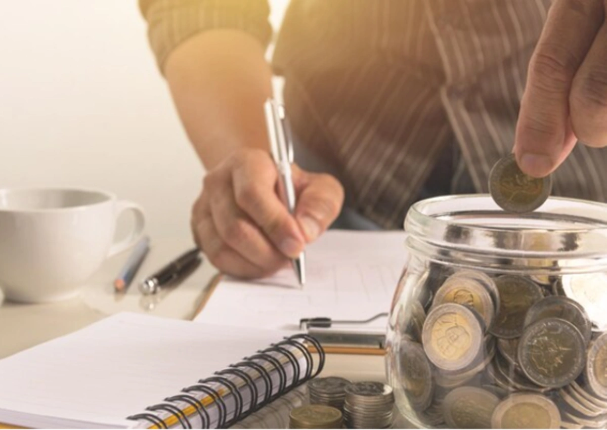Imagem de uma mesa onde um homem está realizando contas, ele escreve algo em um caderno enquanto coloco uma moeda em um cofrinho. Sobre a mesa é possível ver cadernos, canetas, moedas e uma xícara.