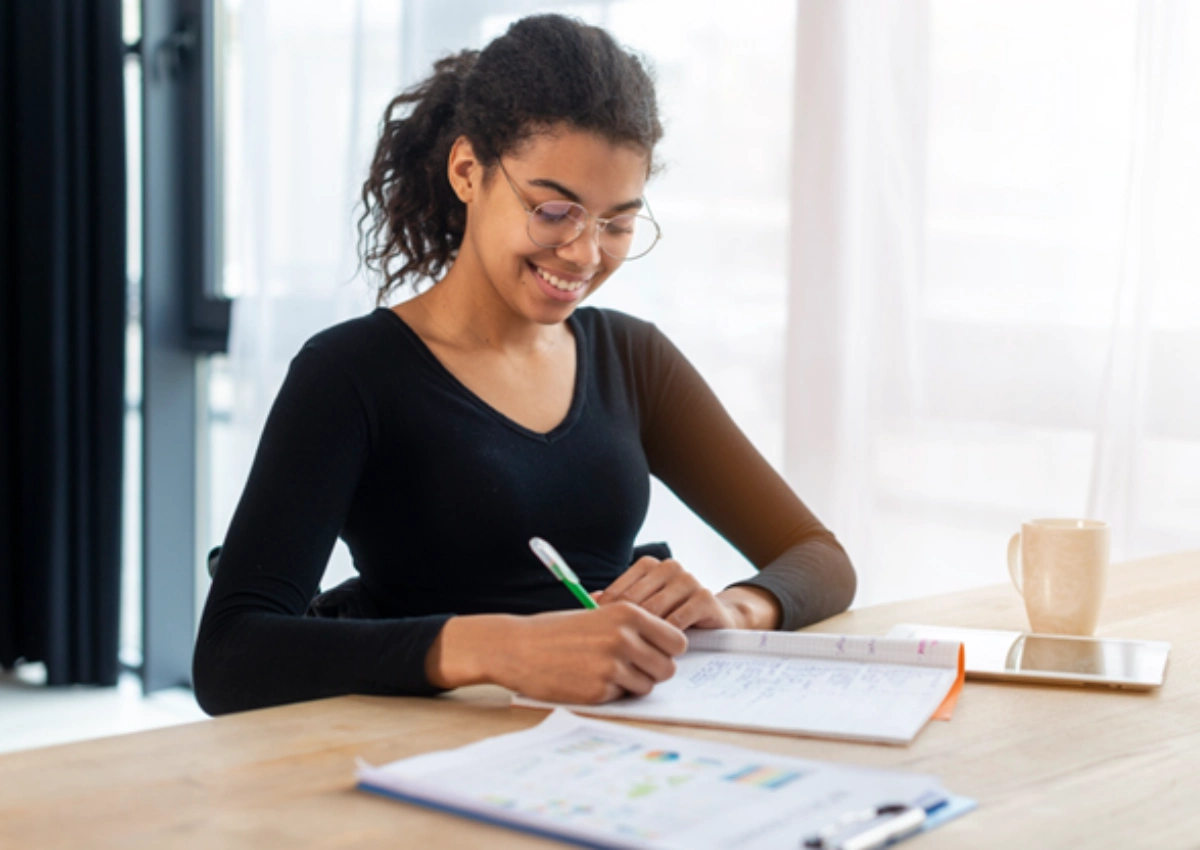 Mulher negra fazendo anotações de caneta no caderno, sobre uma mesa de madeira. Ao redor há uma prancheta, tablet e caneca. No fundo, uma porta de vidro com cortinas.