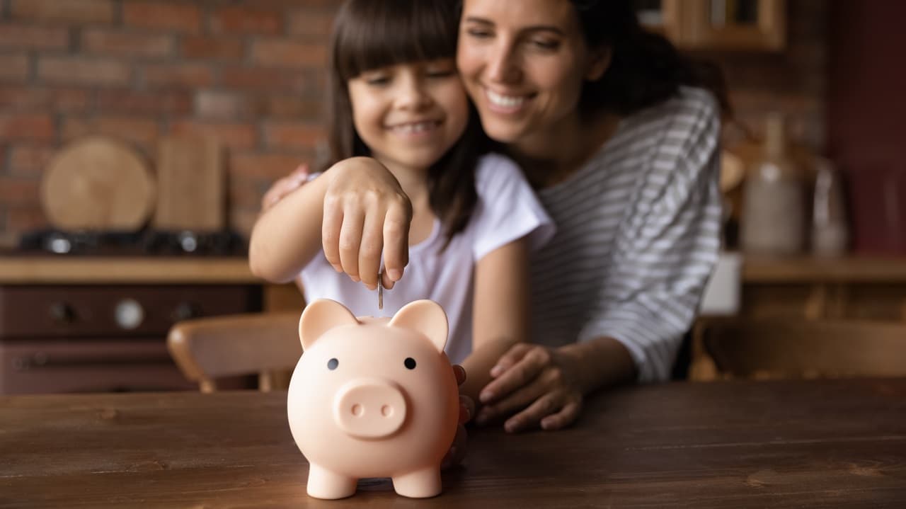  Criança colocando uma moeda em um cofrinho sobre uma mesa, representando poupança e planejamento financeiro