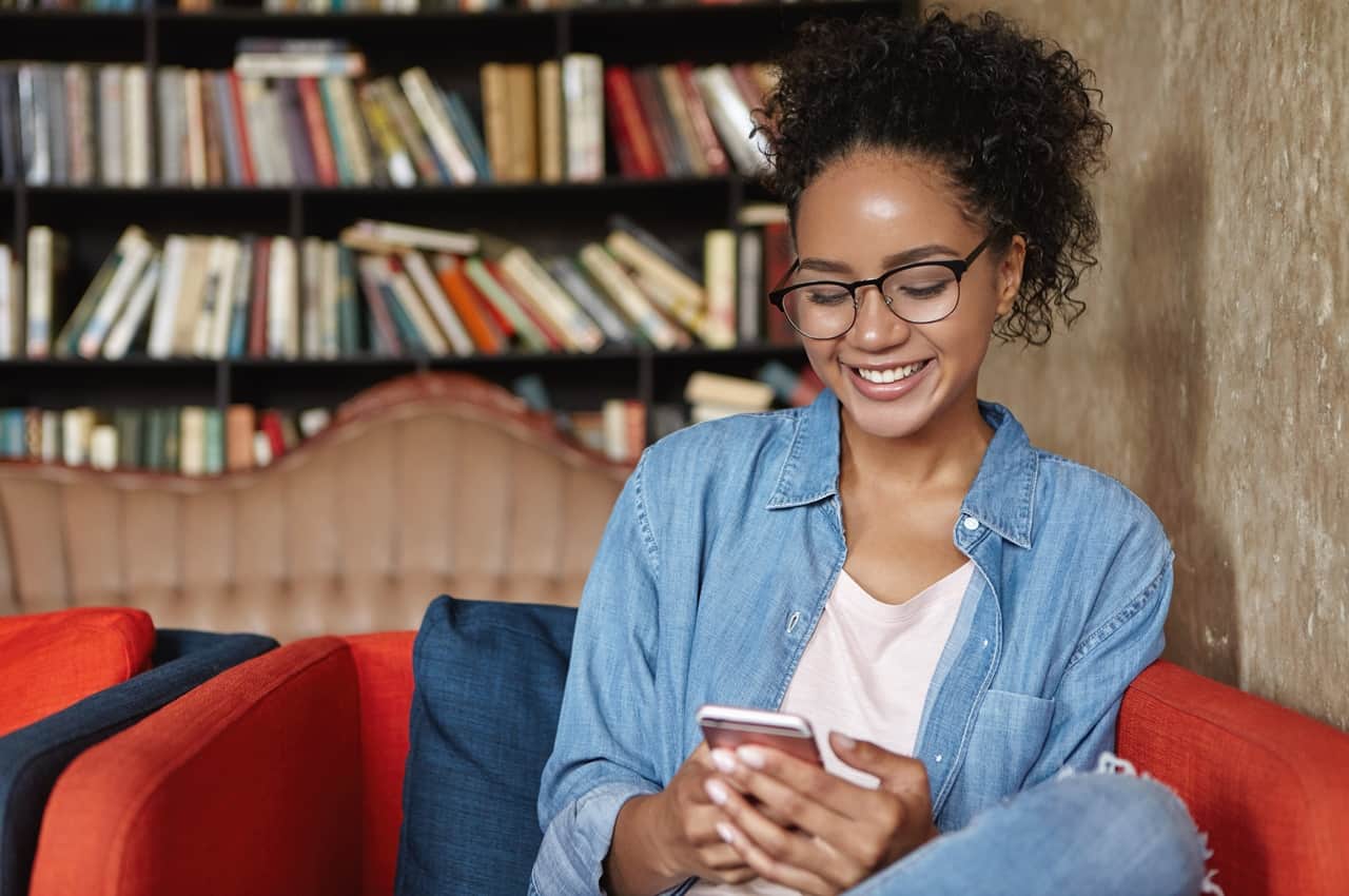 Jovem de óculos e camisa jeans sorrindo enquanto olha para o celular. Ela está sentada em um sofá vermelho com uma estante de livros ao fundo.