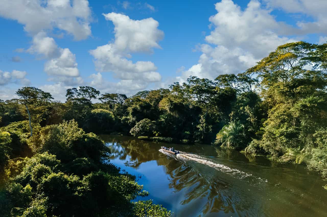 Barco navegando por um rio cercado por vegetação densa, sob céu com nuvens.