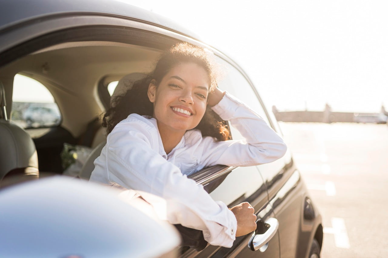 Mulher jovem sorridente debruçada na janela aberta de um carro sob a luz do sol.